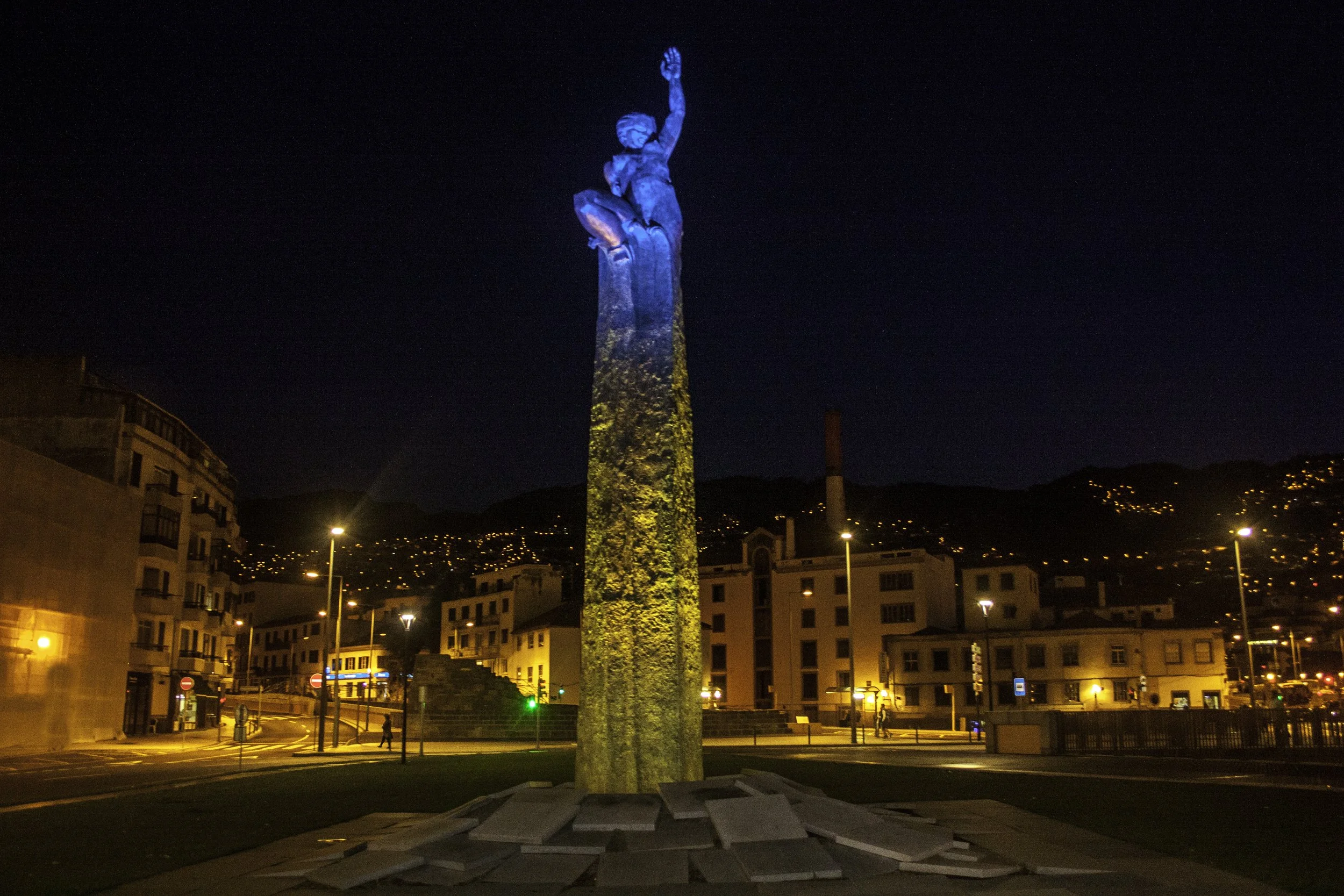 Night view of a statue of a woman sitting on a tall pedestal, with one arm raised, illuminated by colored lights, in an urban area with buildings, streetlights, and hills in the background.
