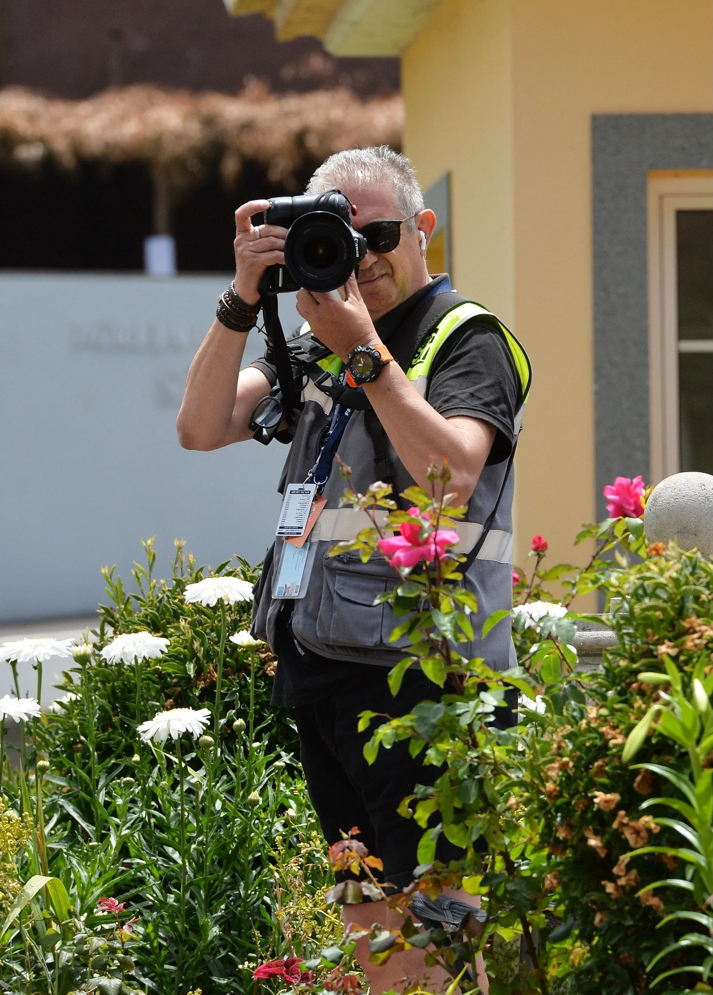 A man with gray hair, sunglasses, and a black shirt stands outdoors among colorful flowers, holding a professional camera and taking photos. He wears a gray vest and has a watch on his left wrist.
