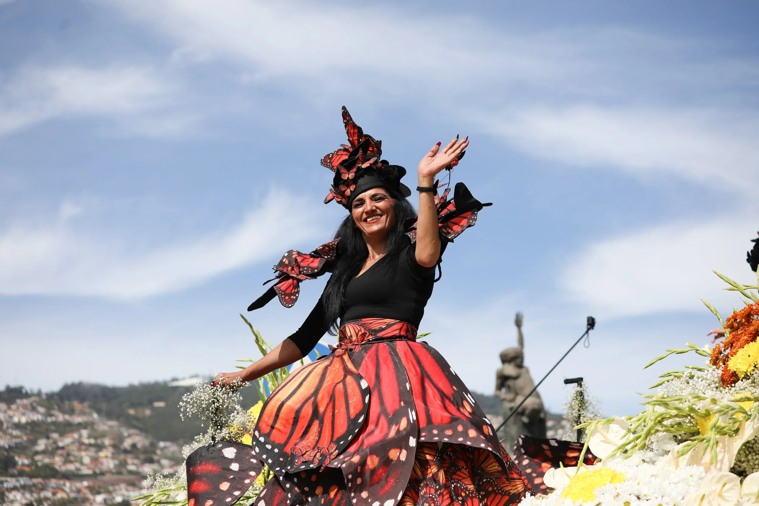 A woman dressed in a butterfly-inspired costume with black and red patterns, smiling and waving during a parade or festival, surrounded by flowers and a clear blue sky.