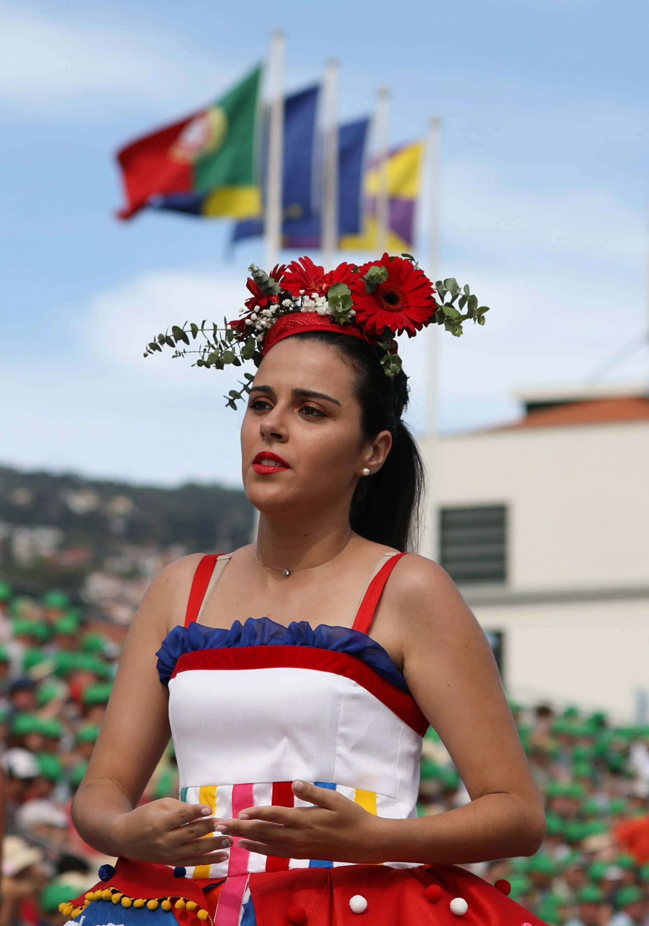 A woman dressed in colorful traditional attire, with a floral headpiece, at a public event or parade with flags in the background.