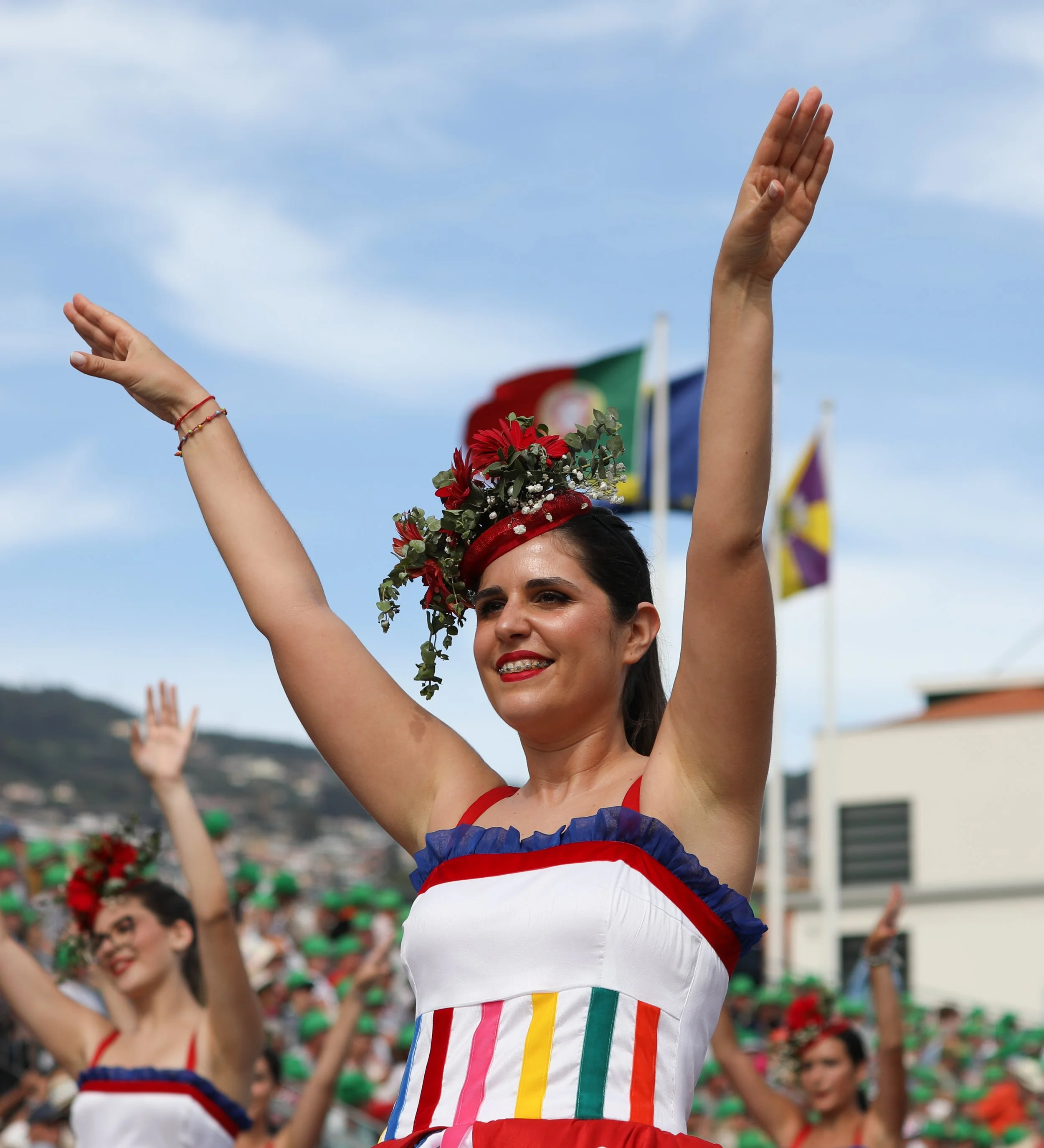 Woman dressed in colorful traditional attire with floral headpiece, smiling, raising arms during a parade or celebration with flags in the background.
