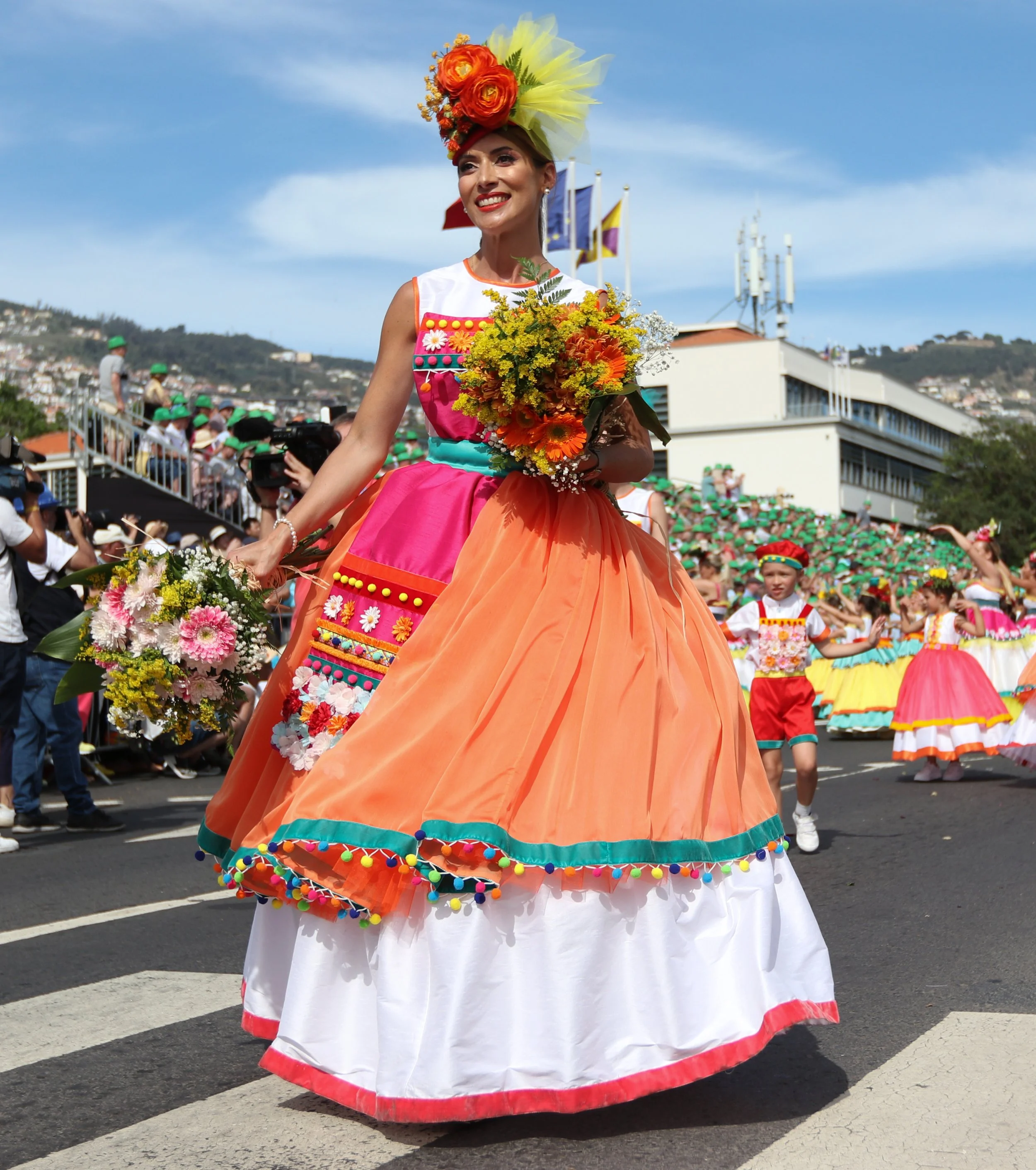 A woman dressed in traditional colorful attire holding a bouquet of flowers during a parade, with children in similar costumes and crowds in the background.