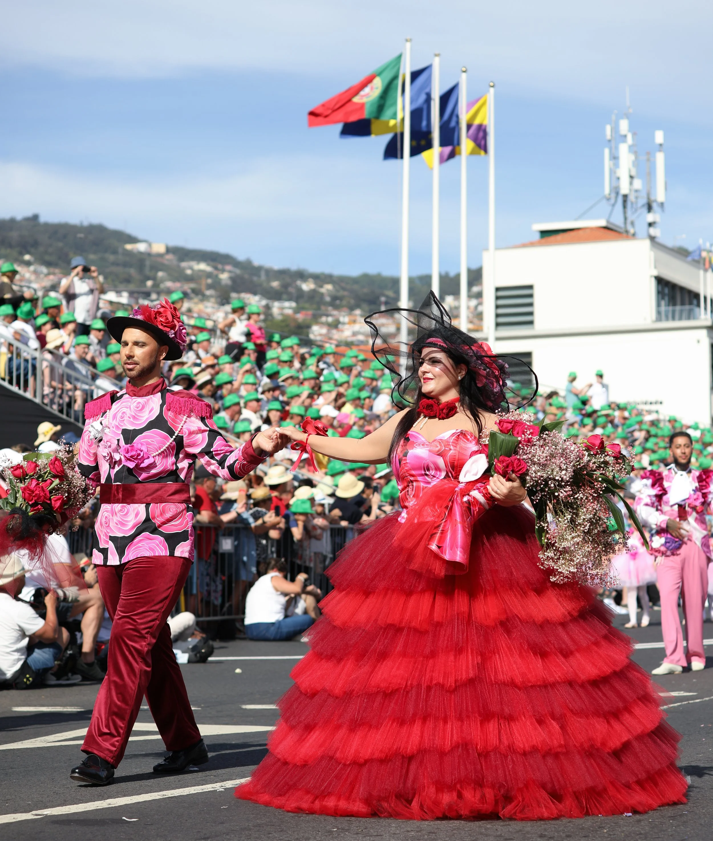 Couple in colorful costumes participating in parade, with audience in background and flags waving