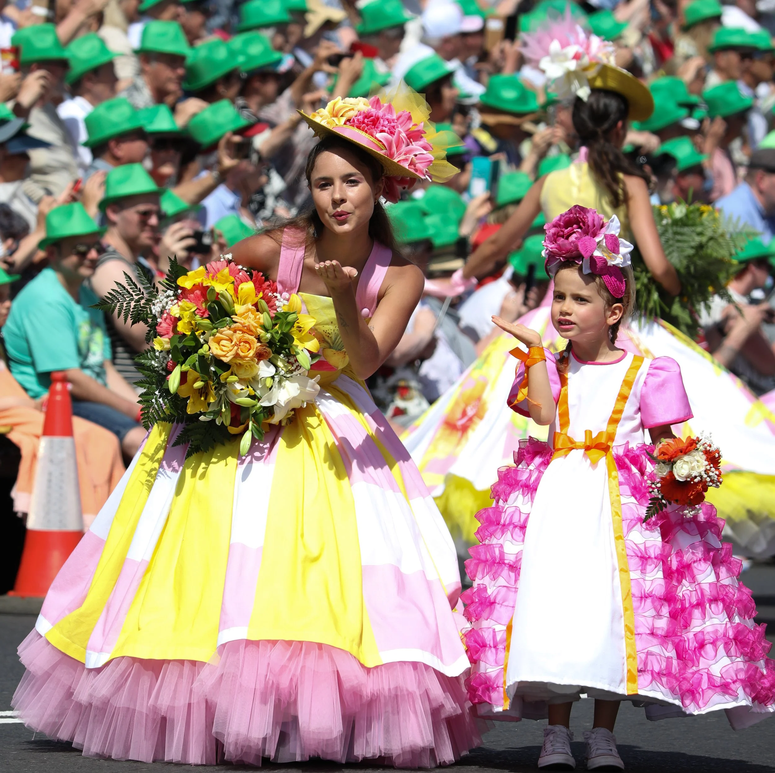Two young girls in colorful, floral dresses participating in a parade, with one blowing a kiss and holding a bouquet of flowers, and the other looking confused or surprised, surrounded by a crowd of spectators wearing green hats.