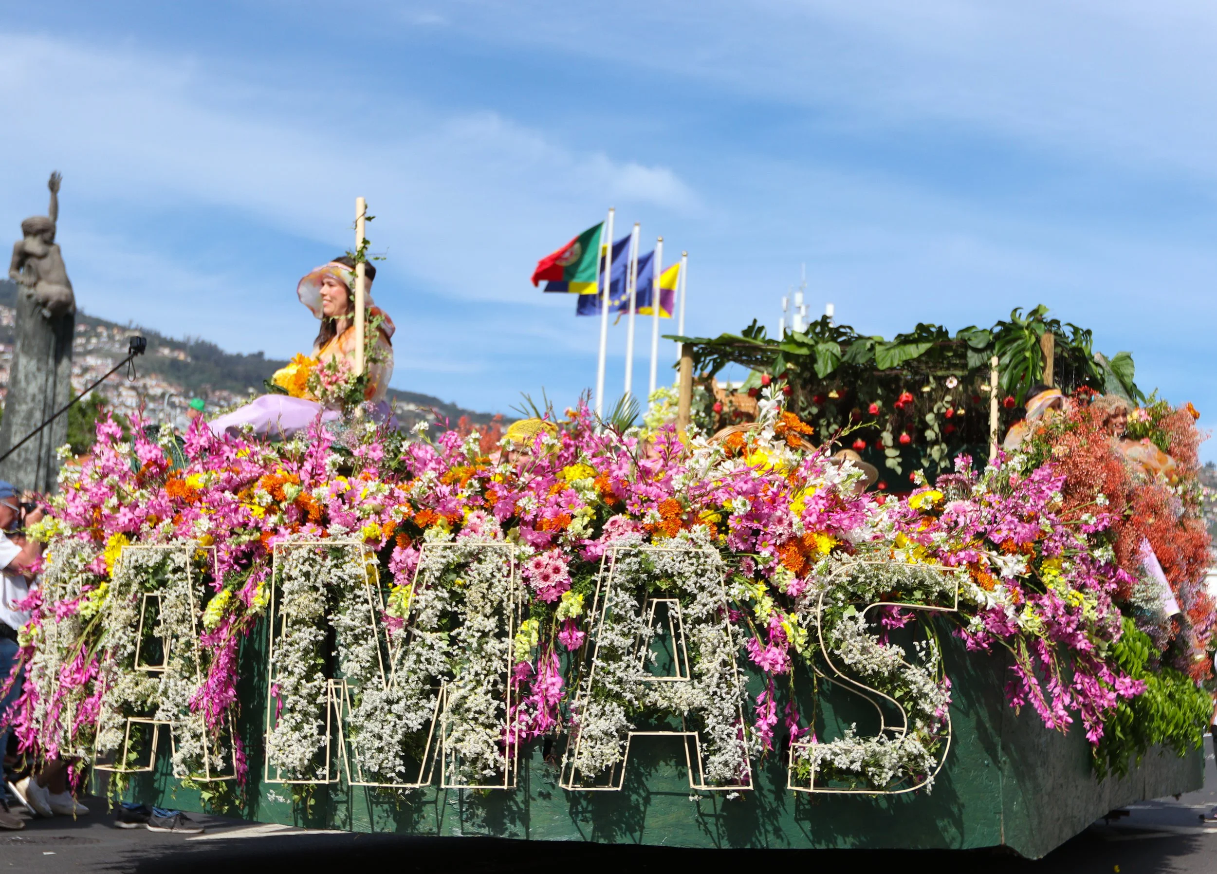 A parade float decorated with colorful flowers and the word 'AVALOS' spelled out in flowers. Two women are sitting on the float, smiling, with a backdrop of flags and a hillside.