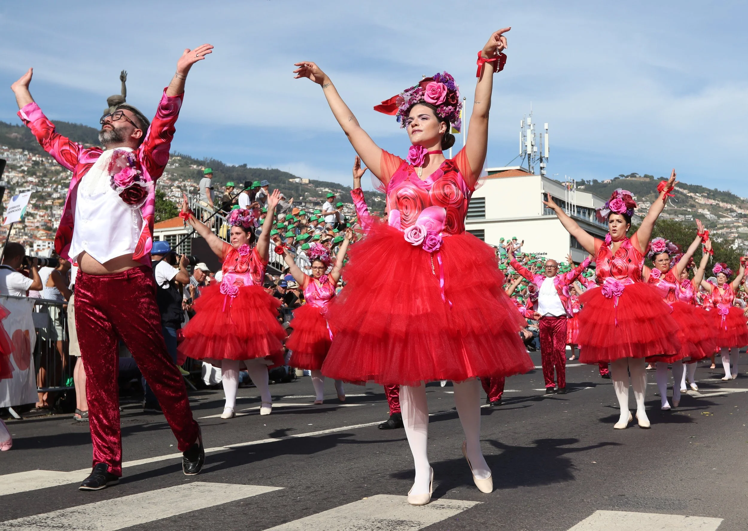 Dancers in colorful costumes performing in a parade, with a woman in a red tutu and pink floral headpiece leading.