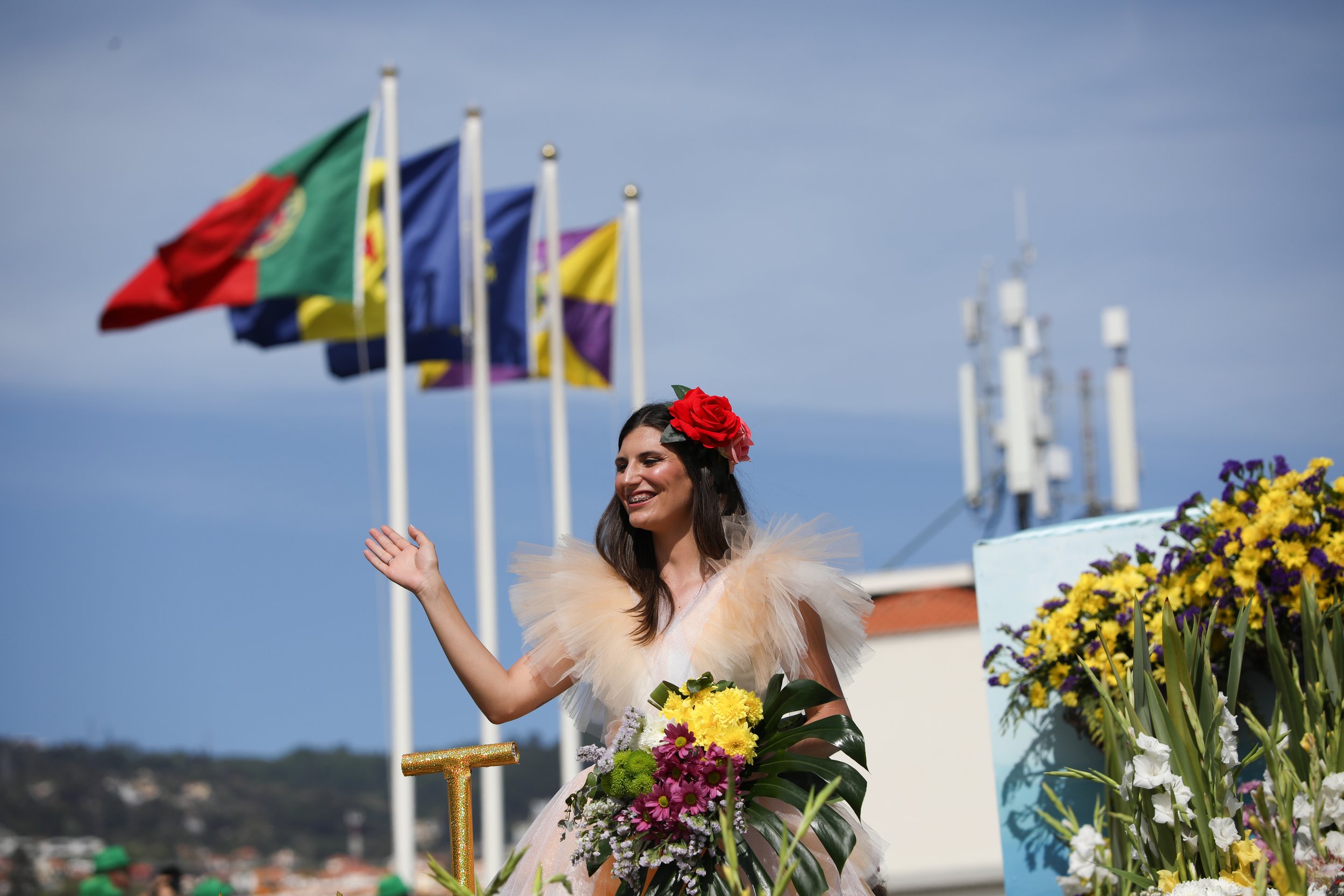 A woman with long dark hair, wearing a dress with tulle ruffles on the shoulders and a large red flower in her hair, holding a bouquet of yellow, purple, and white flowers, and waving. Several flags are flying in the background along with a blue sky 