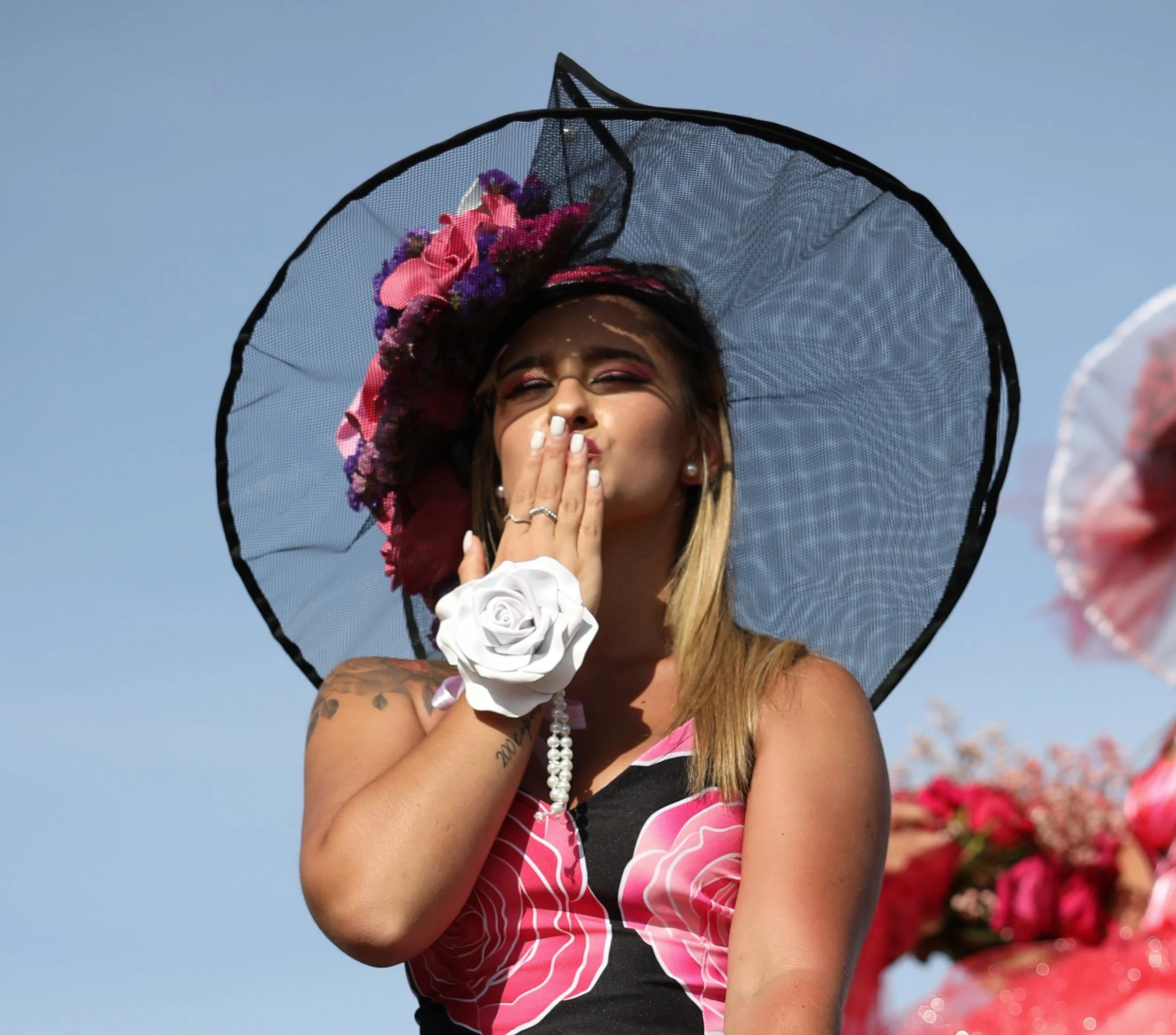 A woman wearing a large, black, see-through hat adorned with pink and purple flowers blowing a kiss, with a tattoo on her arm and a white floral bracelet, against a clear blue sky.
