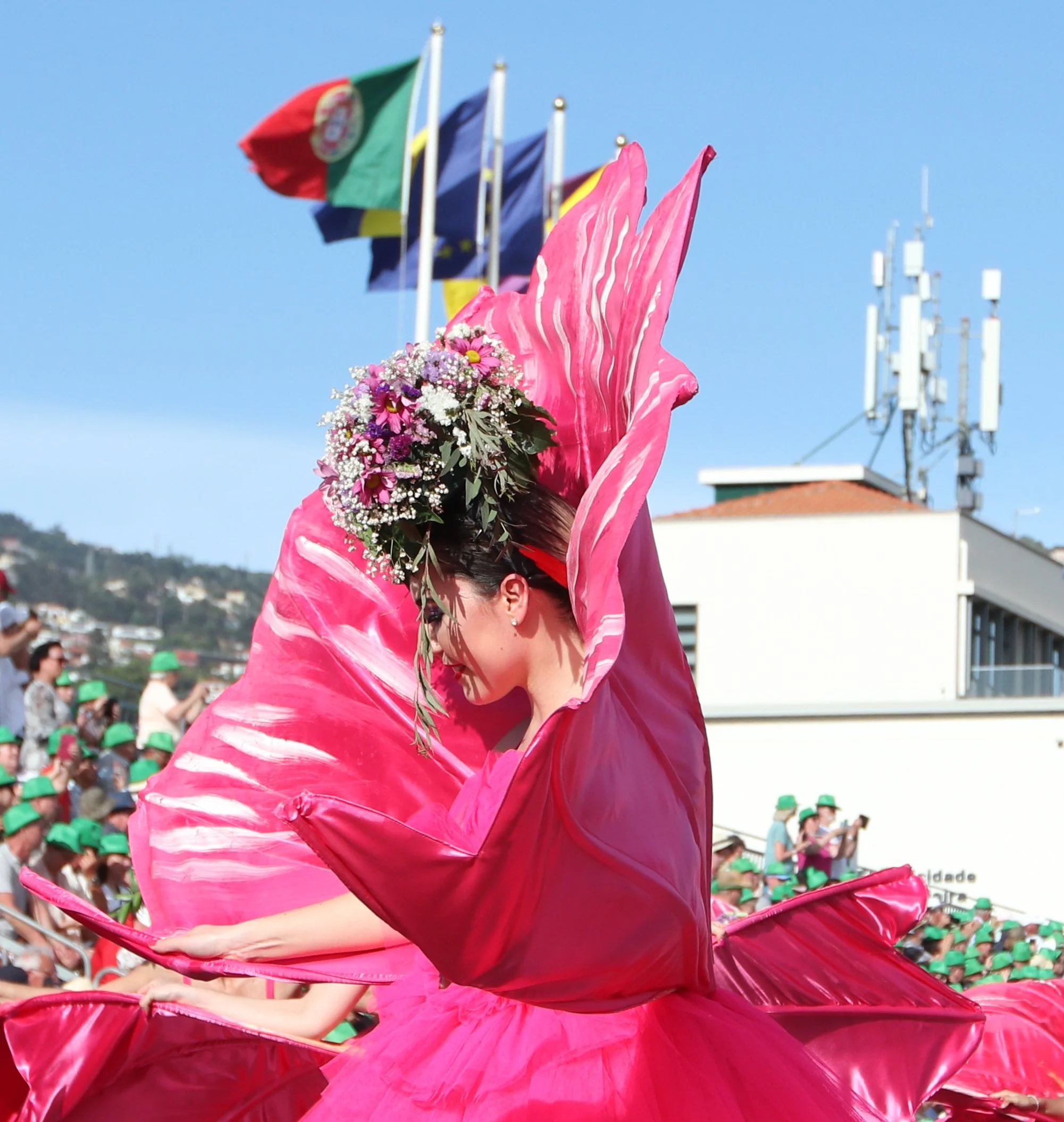 A woman in a vibrant pink floral costume dancing at a parade, adorned with a floral headpiece, under a clear blue sky, with flags and a crowd in green hats in the background.
