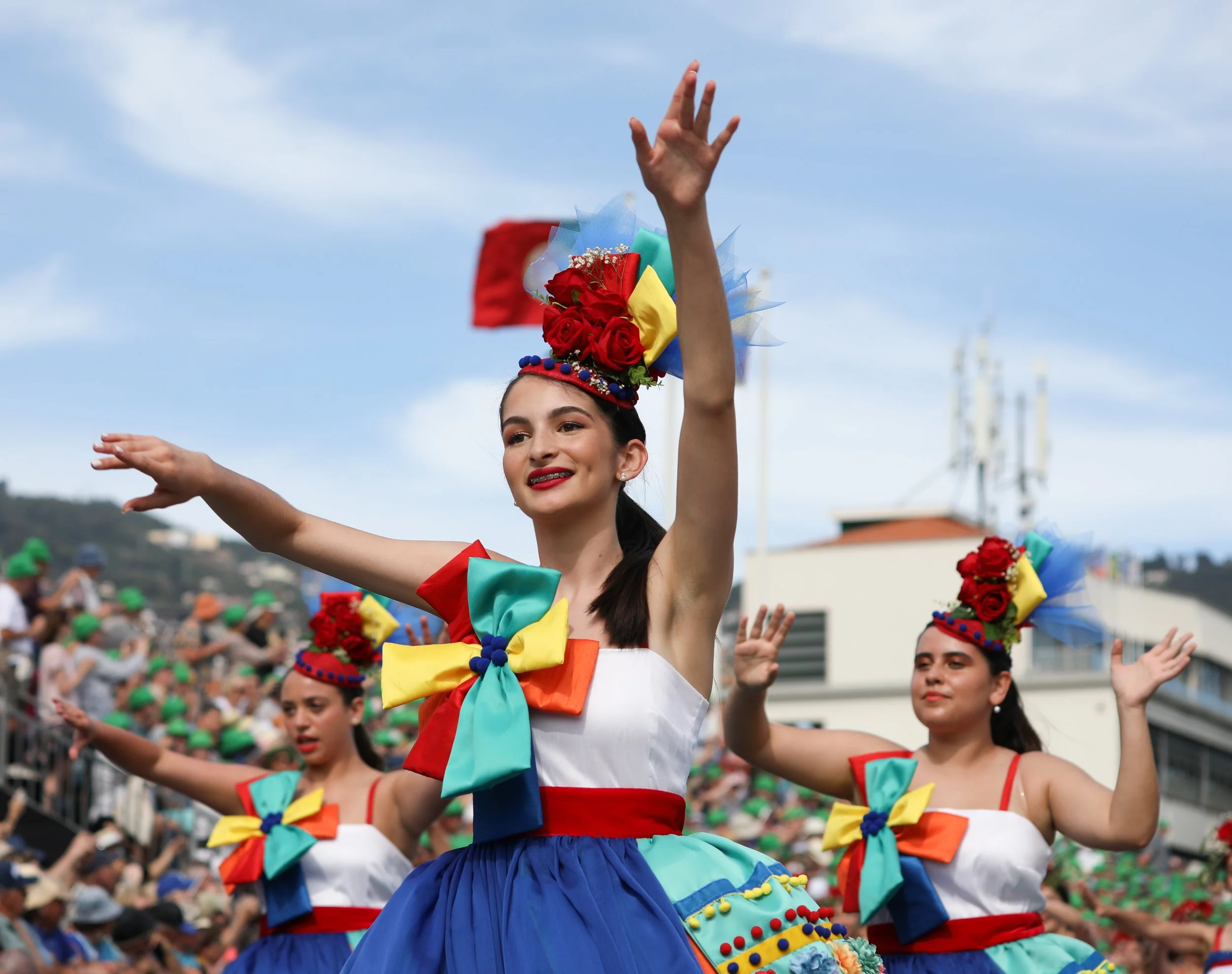 Young women dressed in colorful traditional costumes, dancing at a parade with a crowd and cityscape in the background.