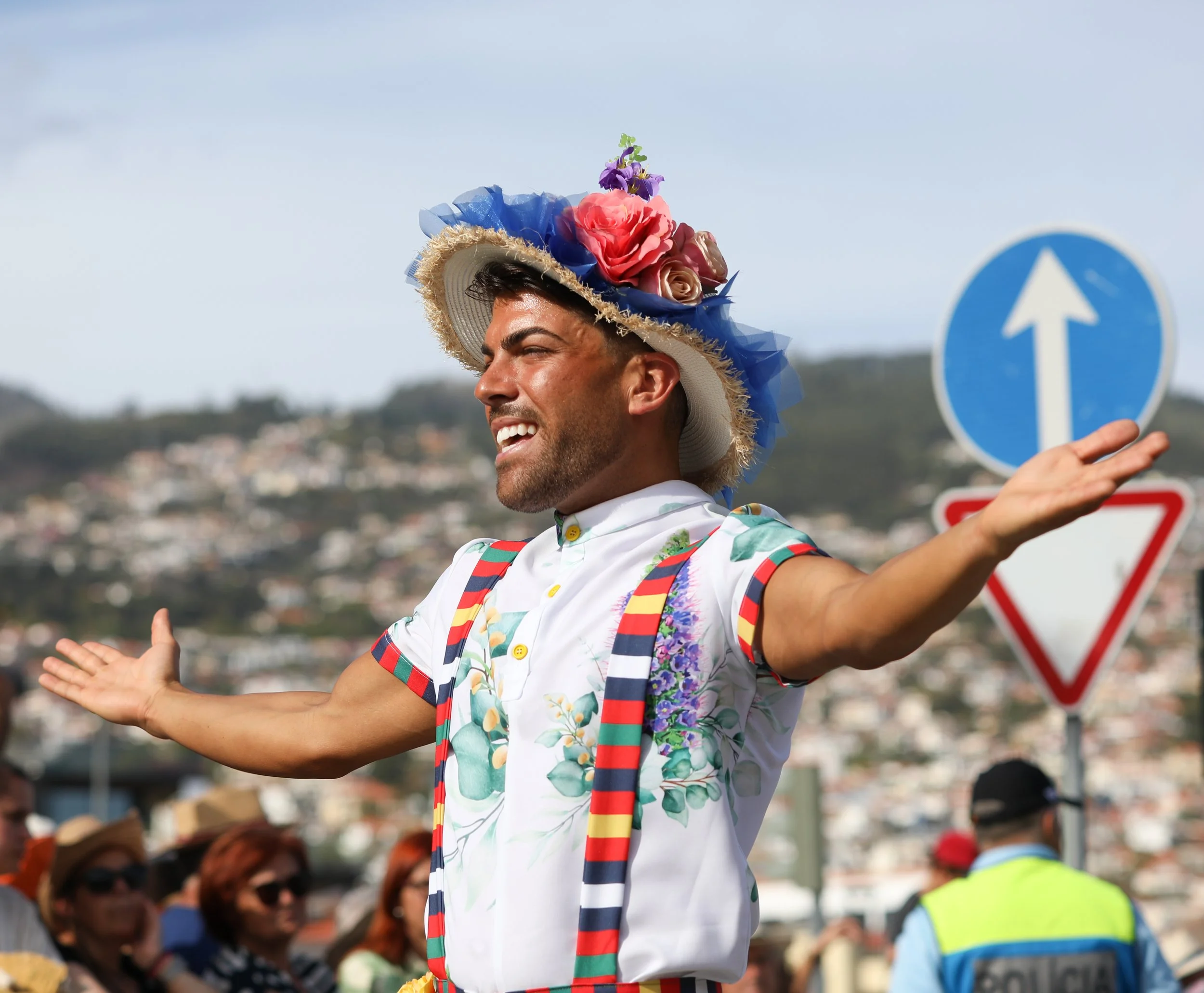 A man wearing a floral shirt, striped suspenders, and a large hat decorated with colorful flowers, standing outdoors with his arms open, smiling, with a crowd in the background and traffic signs nearby.