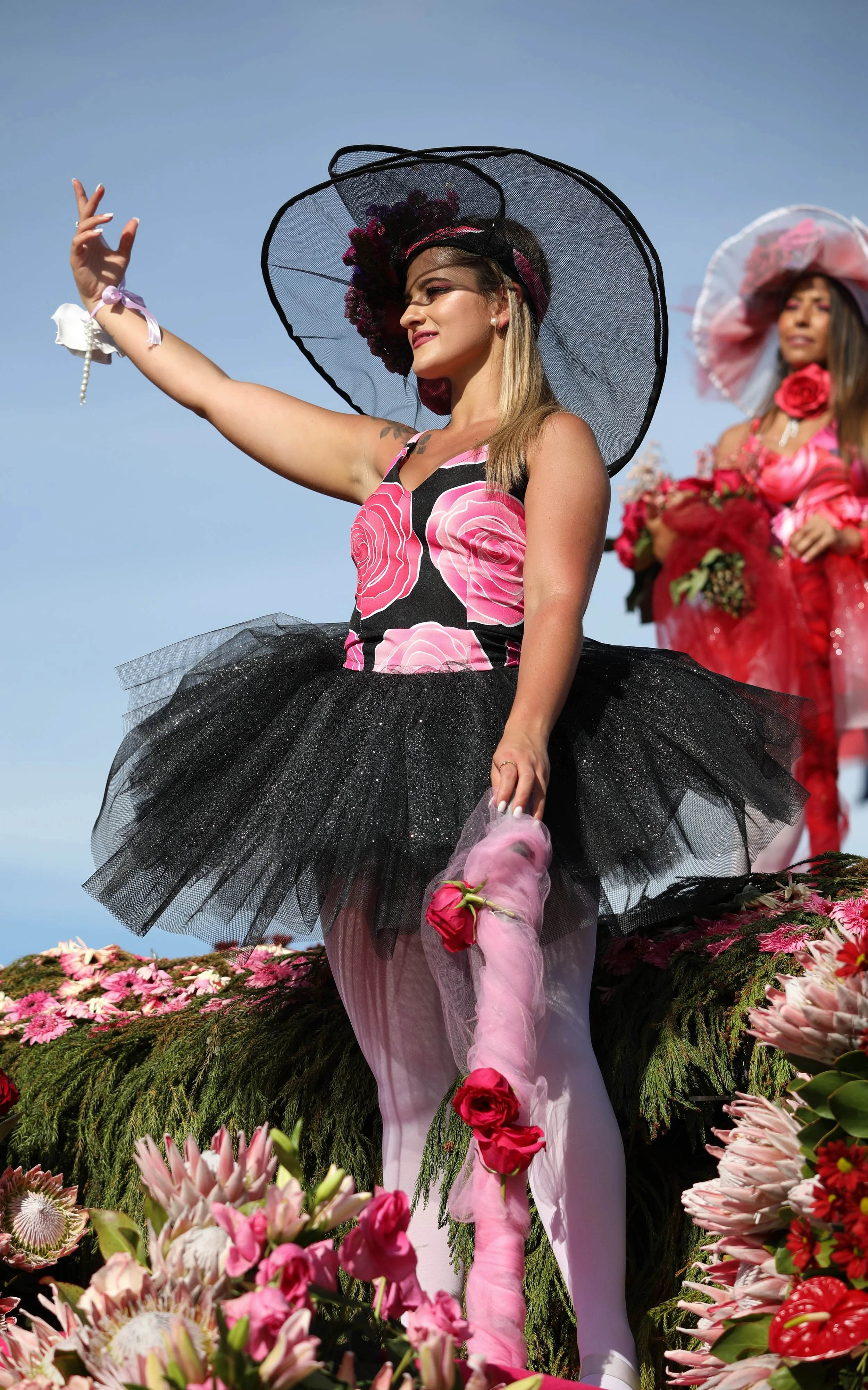 A woman in a floral-themed outfit with a black tutu, wearing a large black hat with pink flowers, holding a pink flowered staff, standing on a float surrounded by pink and red flowers.