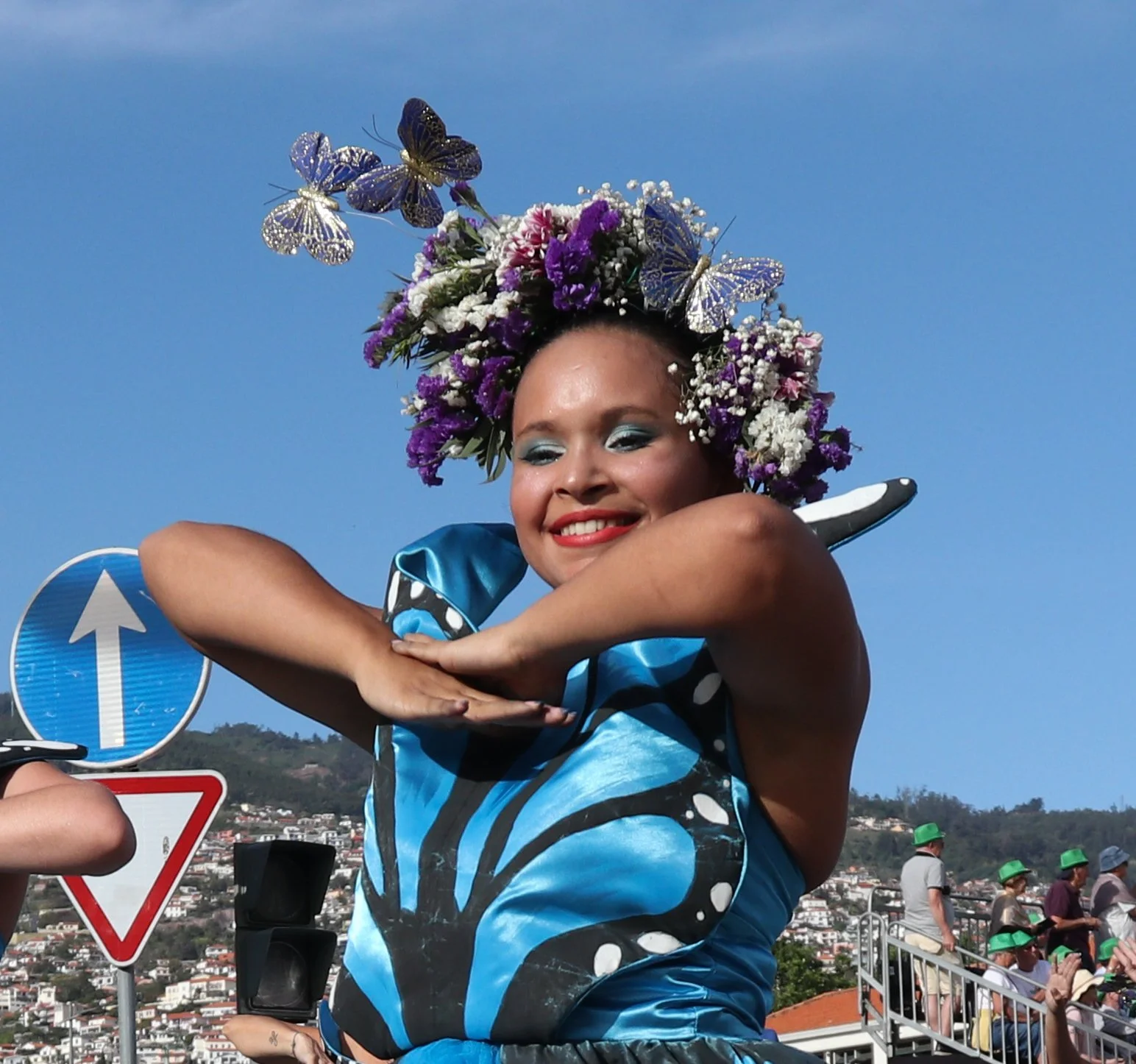 A woman wearing a blue dress with butterfly patterns and a headdress made of colorful flowers and butterflies, smiling with arms crossed near a street with traffic signs and a crowd in the background.