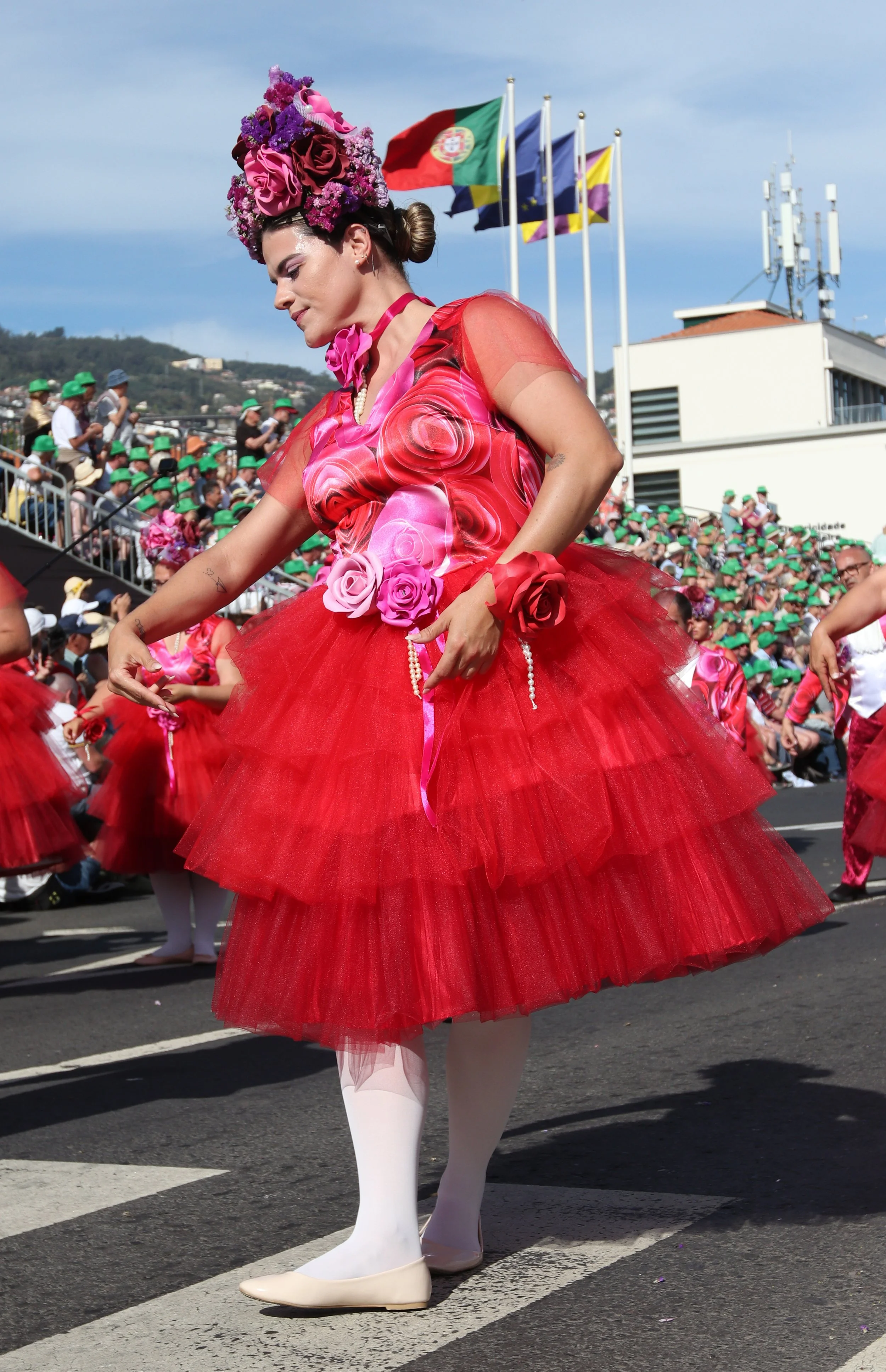 A woman in a colorful costume with a floral headpiece, red Tulle skirt, and white shoes participating in a parade with flags and spectators in the background.