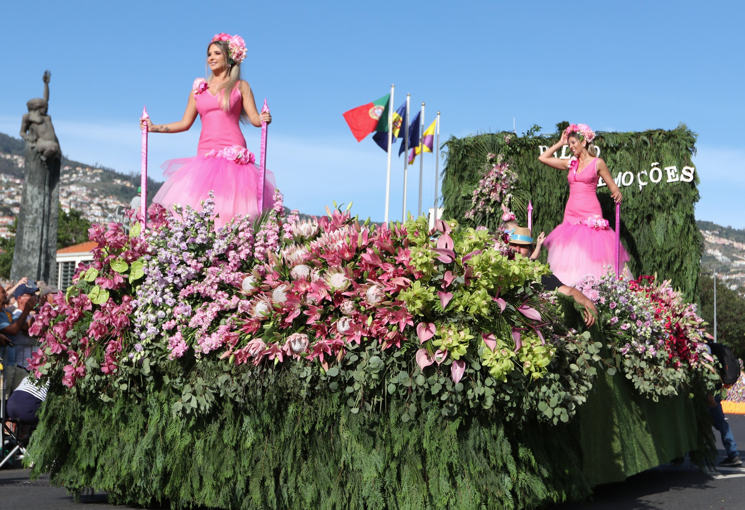 A parade float decorated with pink flowers, featuring two women in pink dresses and floral crowns. The float is outdoors with flags and a city hillside in the background.