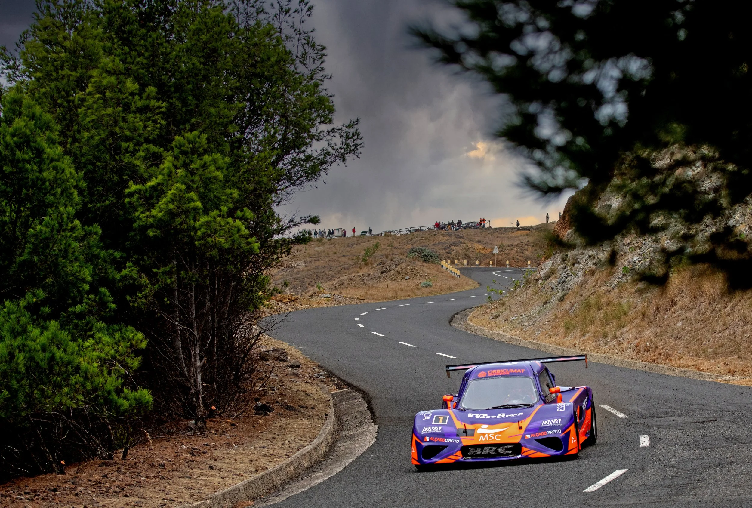 A race car driving on a winding mountain road with spectators in the distance and trees on the side.