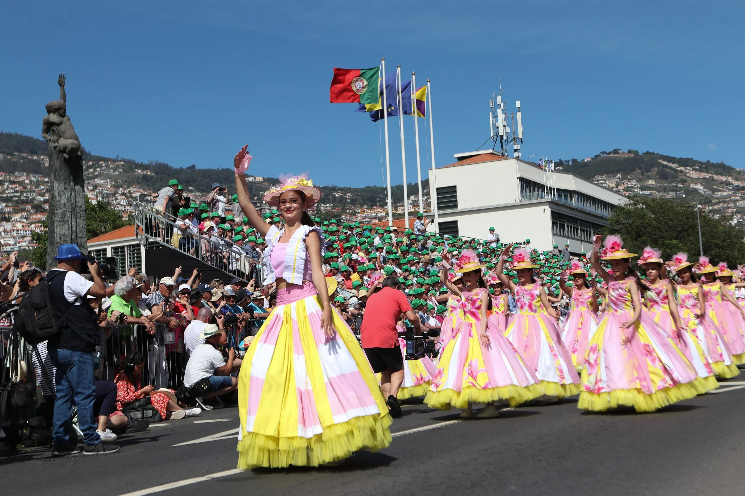 People parade with women in pink and yellow dresses, some with large hats, waving to the crowd during a celebration on a sunny day with clear blue sky.