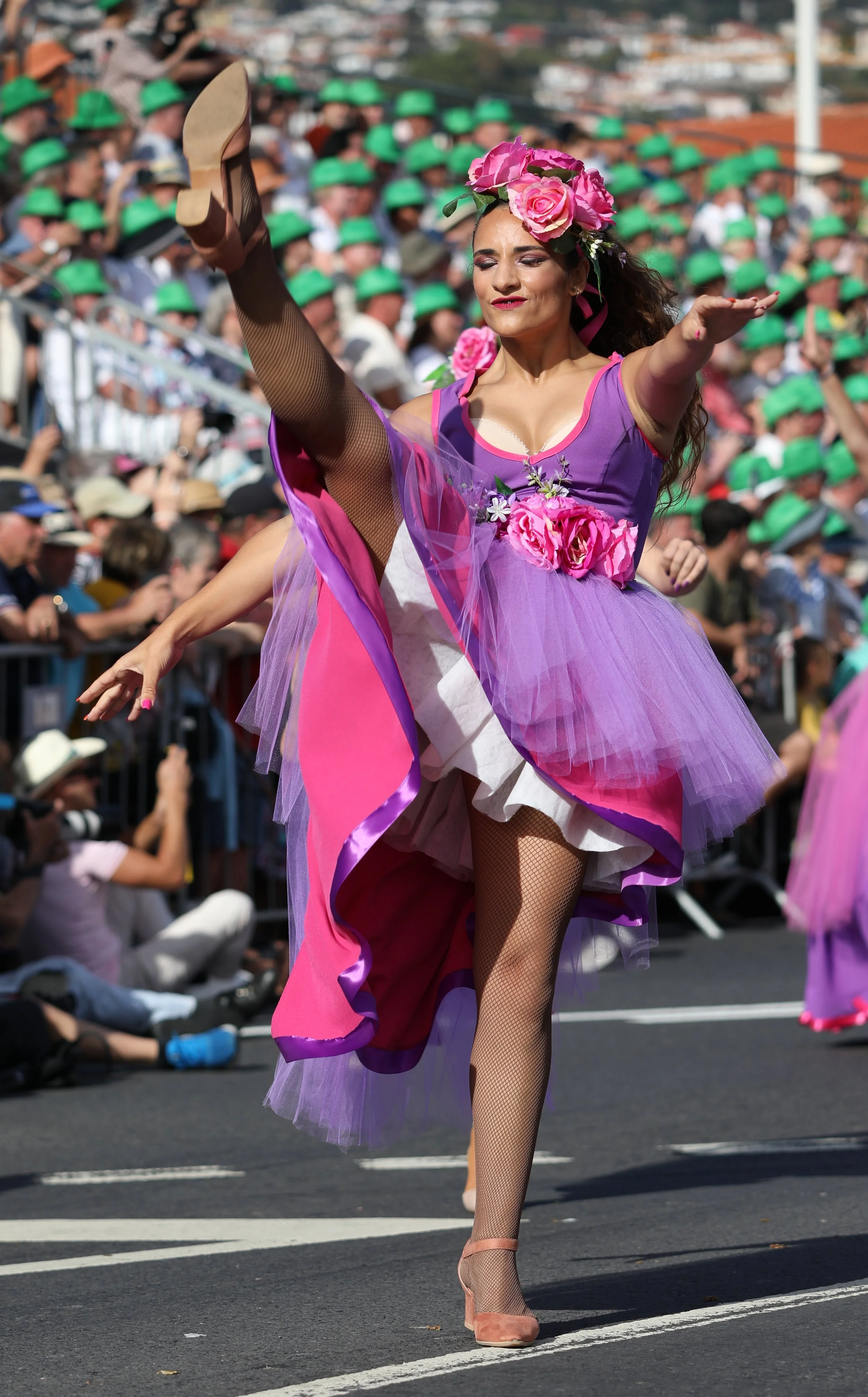 A woman in a colorful costume performing a dance during a parade, with a large crowd of spectators in the background.