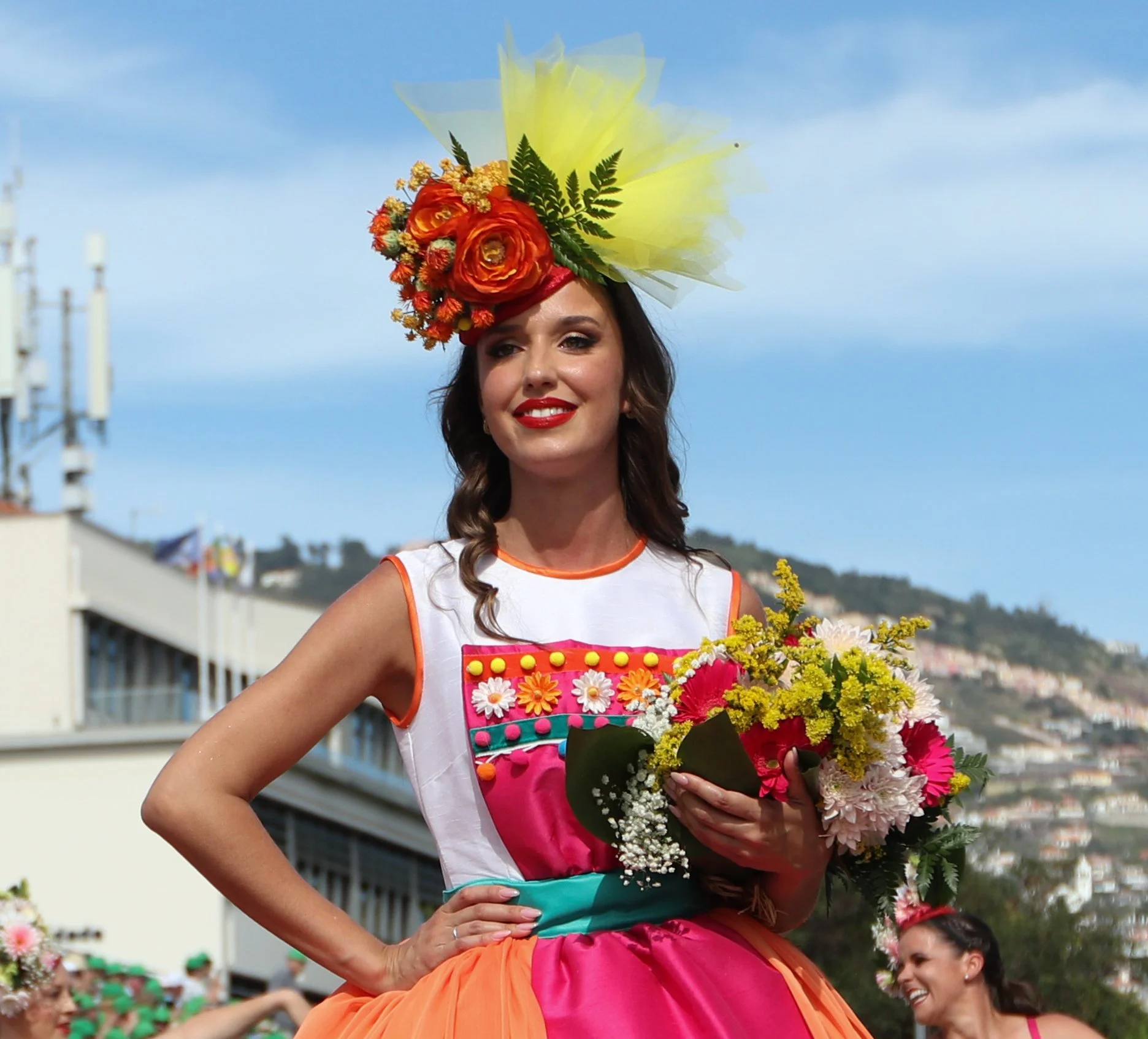 Young woman in colorful dress with a floral headpiece and bouquet, standing outdoors during daytime.