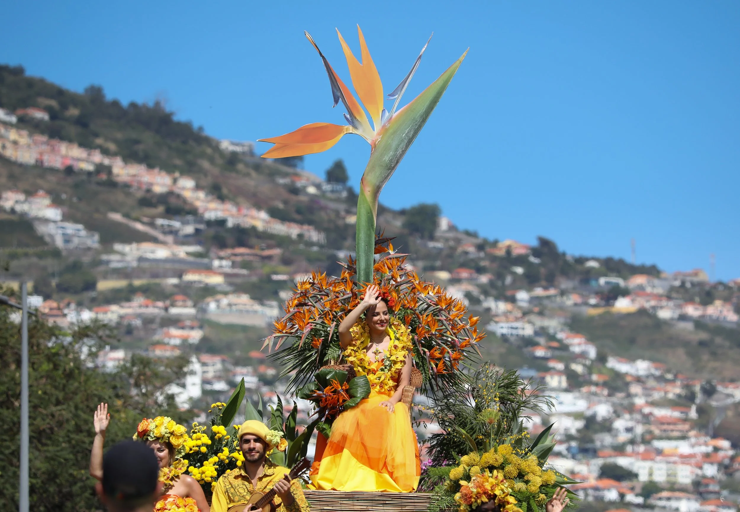 A parade float decorated with tropical flowers and greenery featuring a woman in a yellow dress and floral lei, waving to spectators against a backdrop of hillside houses and a clear blue sky.