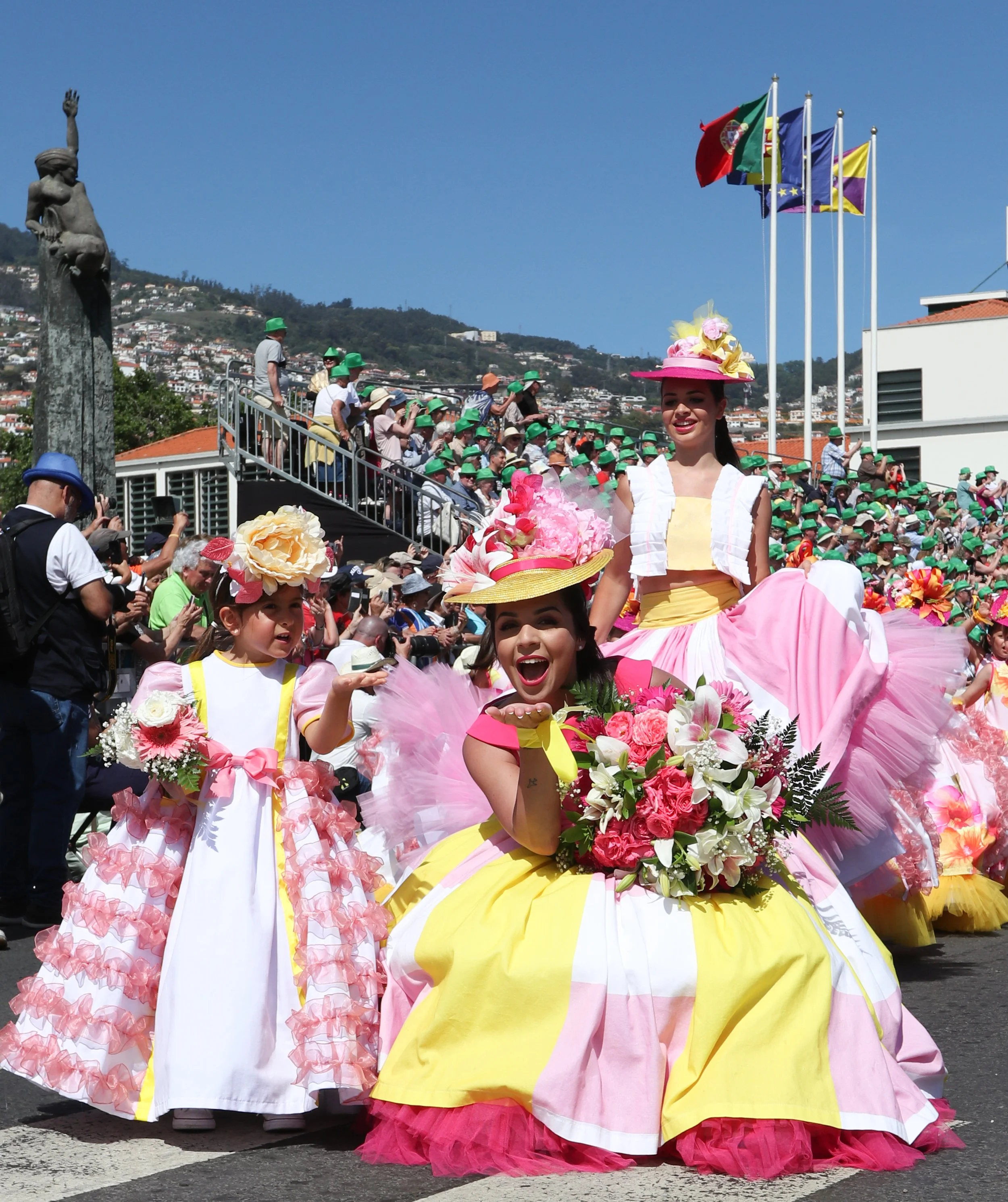 Women and a girl dressed in colorful, traditional dresses participating in a parade with flags and a crowd of spectators in the background.