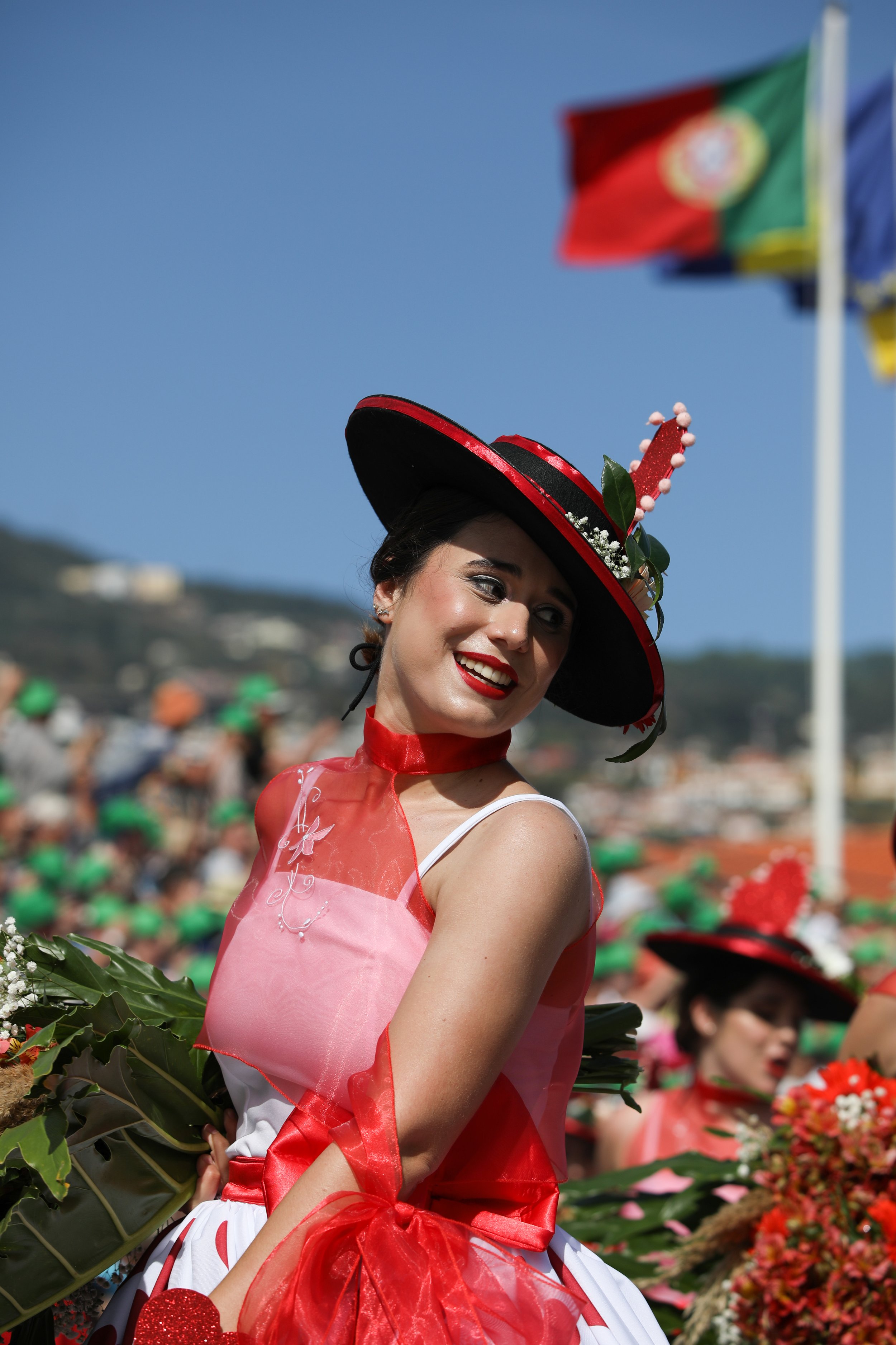A woman smiling in traditional attire at a festival, holding a bouquet of flowers, with flags in the background.