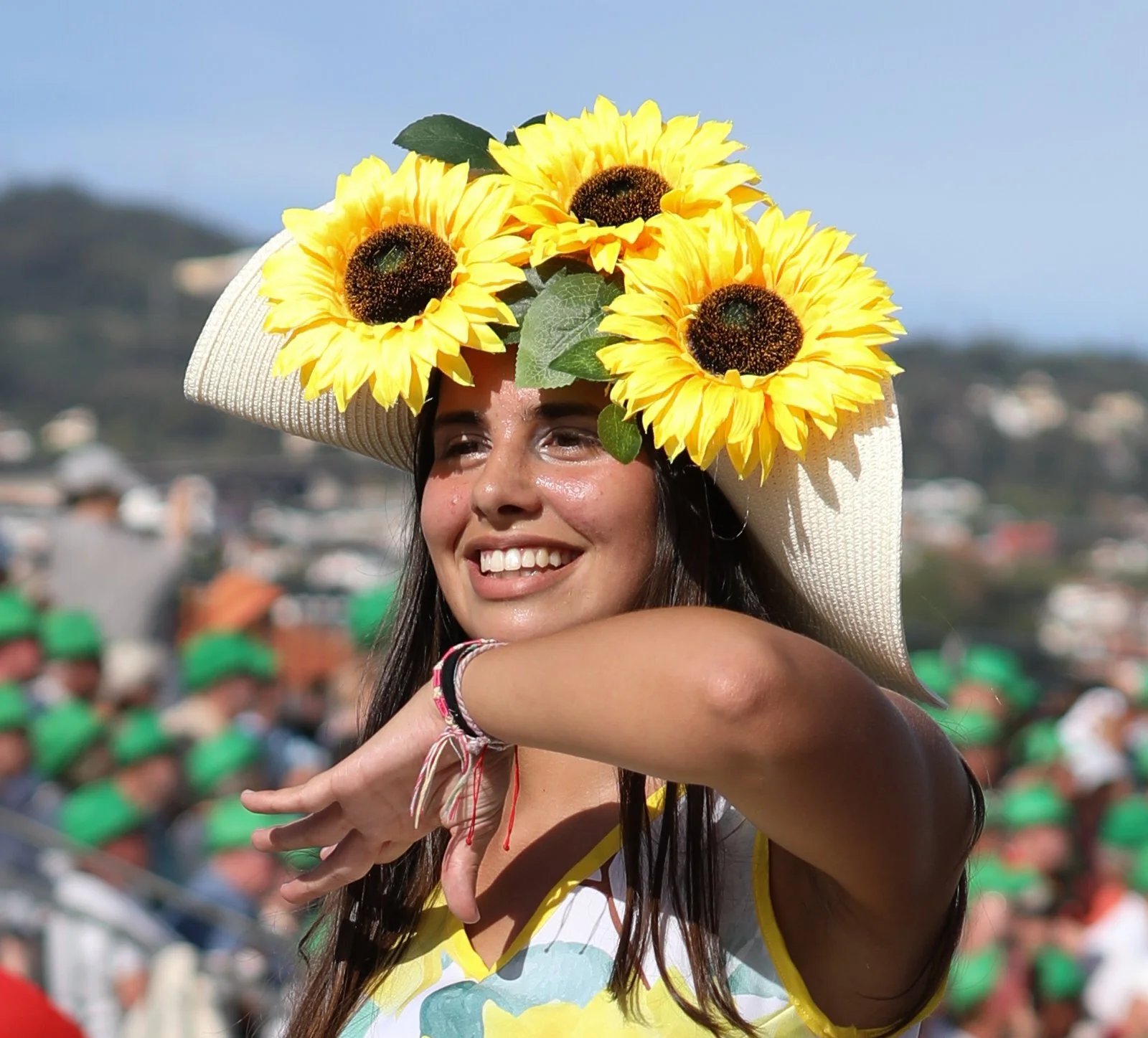 A young woman wearing a large straw hat decorated with sunflowers, smiling and enjoying the outdoors.