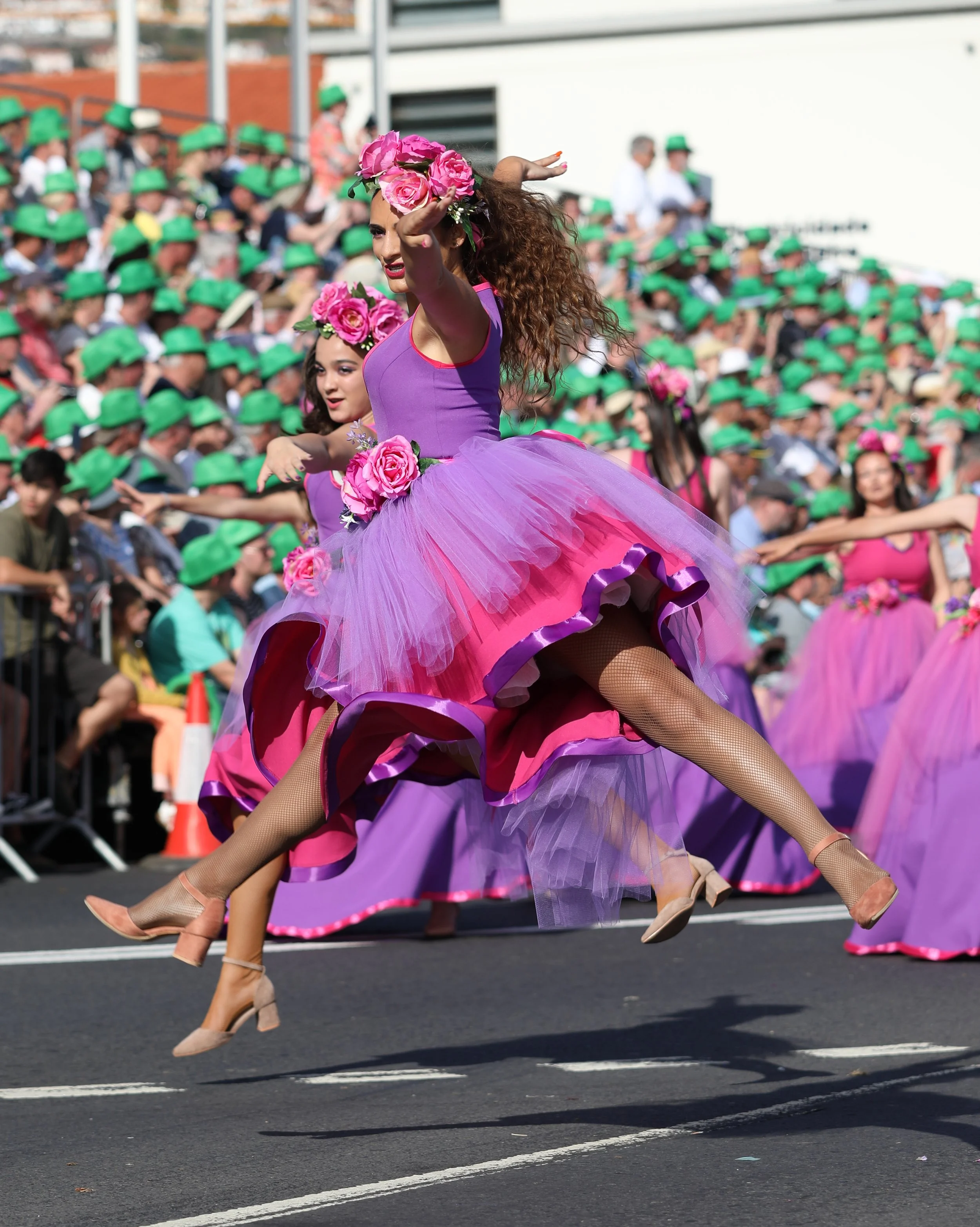 Women wearing vibrant pink and purple flower-adorned dresses perform a dance in a parade, with spectators watching from the stands.