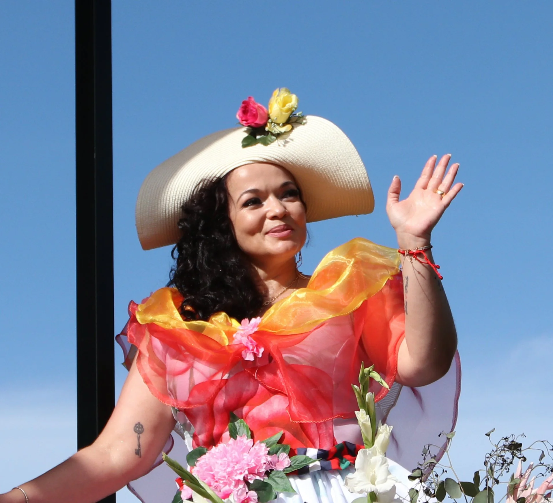 Woman wearing a large wide-brimmed hat with flowers, colorful ruffled dress, holding flowers, and waving outdoors with blue sky