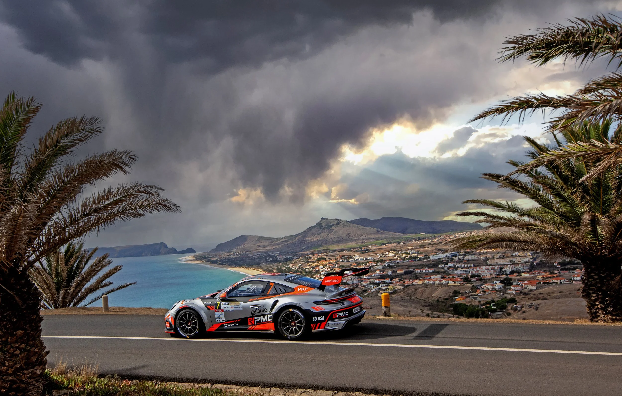 A race car on a curving mountain road with palm trees in the foreground, a coastal town, and mountains in the background under a cloudy sky.