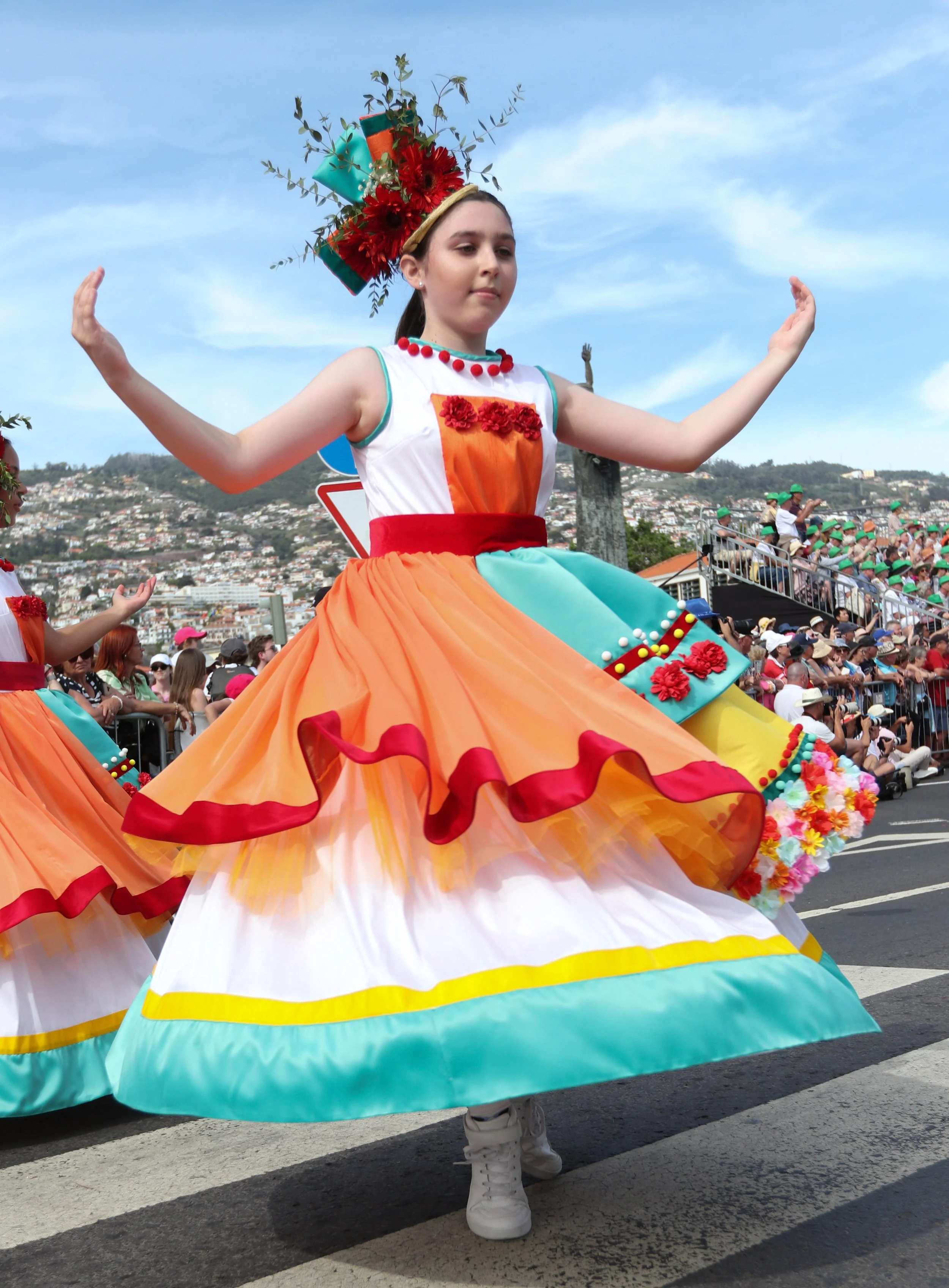 A young woman participating in a parade, wearing a colorful traditional dress with a wide, flowing skirt and a floral headpiece, surrounded by a crowd of spectators.
