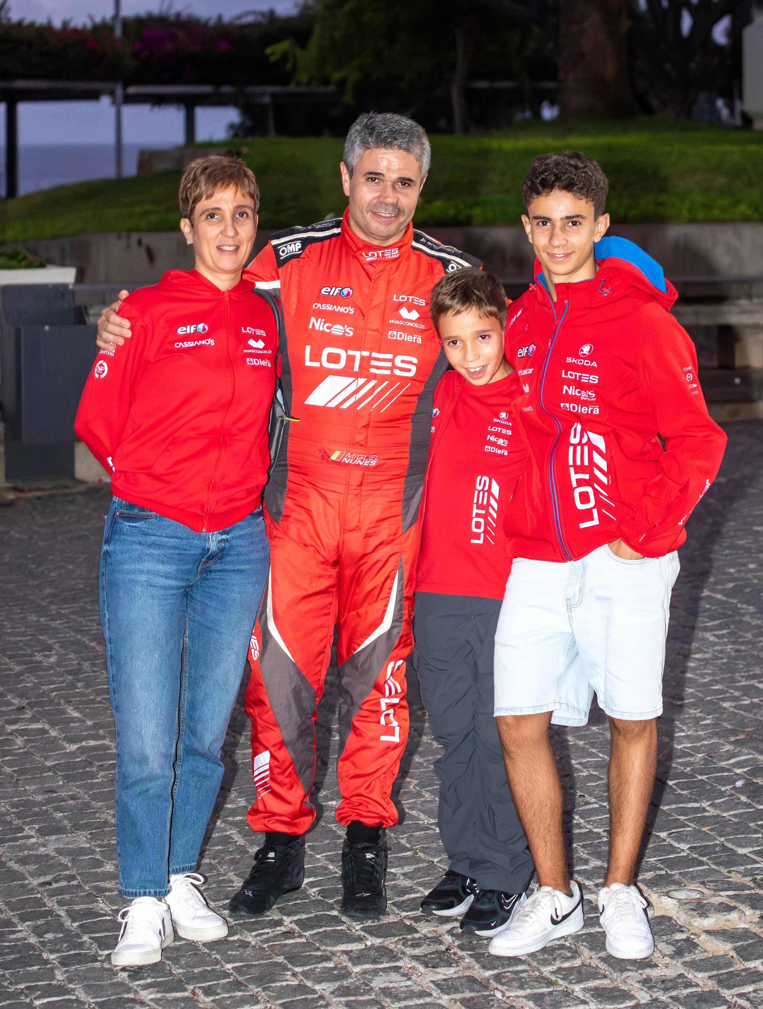 A family of five wearing matching red and black racing team jackets and overalls, standing together outdoors on a cobblestone surface, with a park and trees in the background, smiling for the camera.