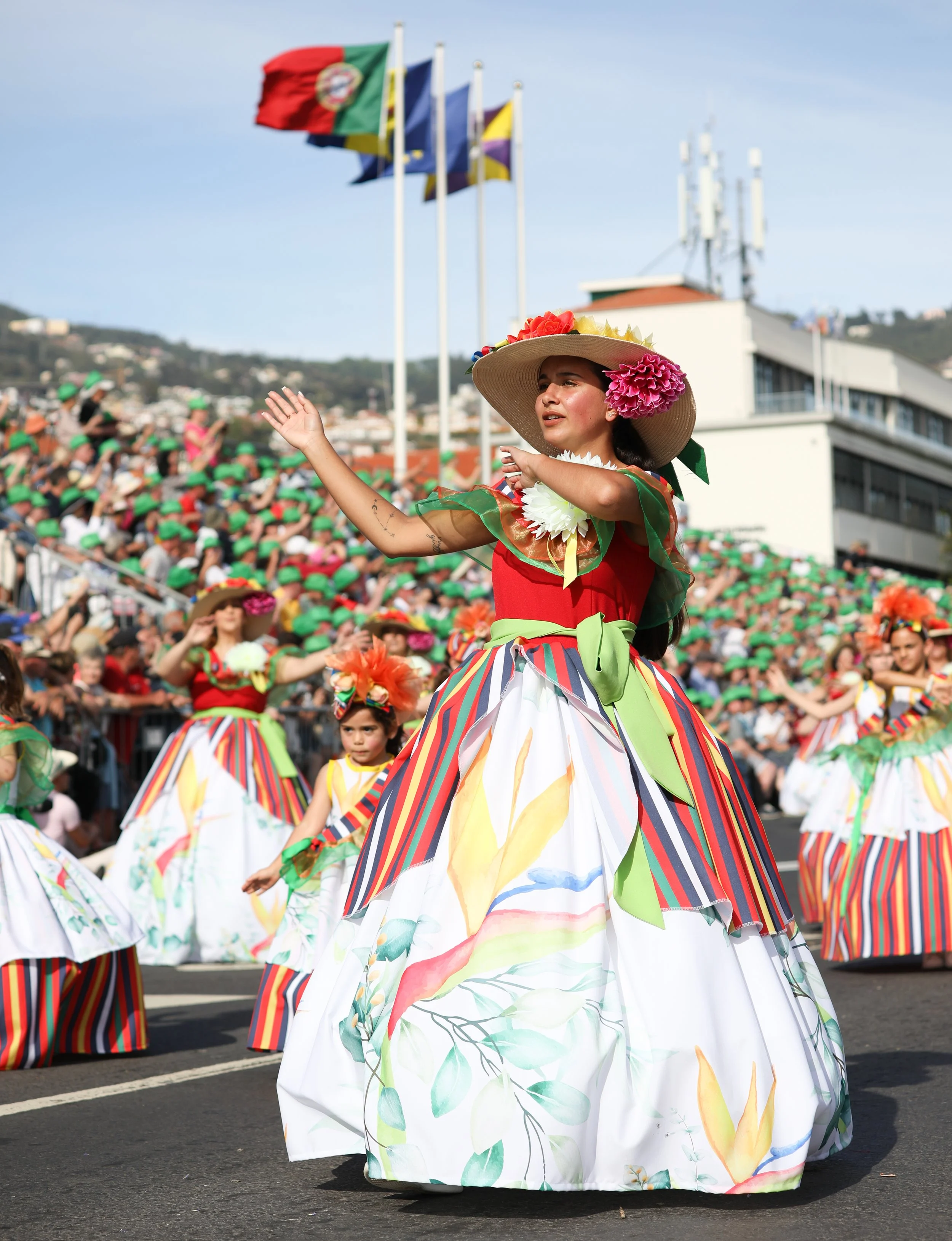 Women and children dressed in colorful traditional dresses and hats participate in a parade on a sunny day with a large crowd and flags in the background.