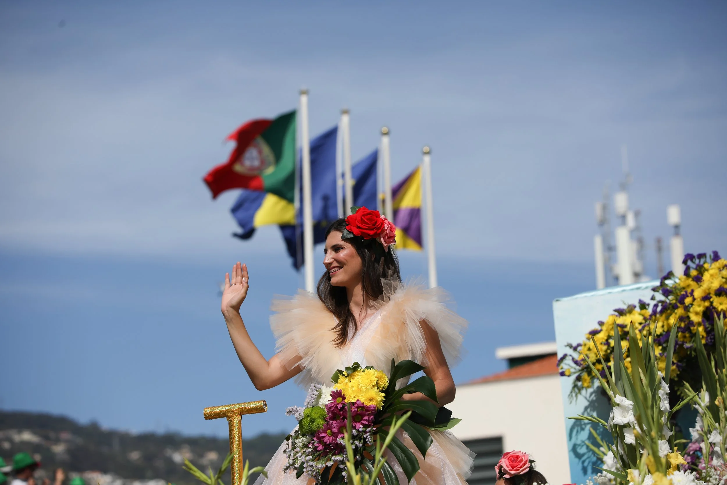 A woman in a fancy dress with tulle ruffles and a floral headpiece, holding a bouquet of flowers, waving at an outdoor event with flags in the background.