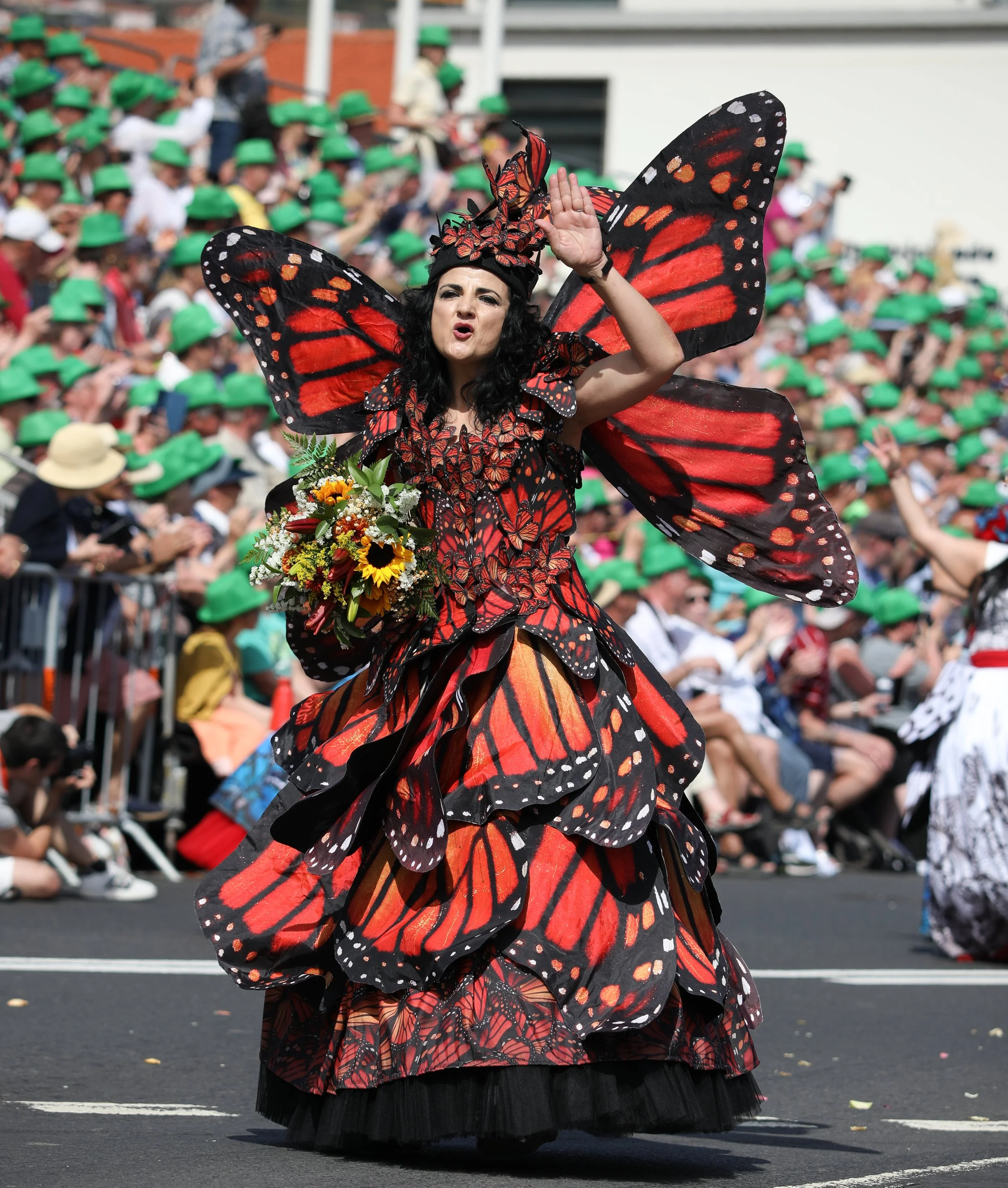A woman dressed as a butterfly with red and black wings participating in a parade, holding a bouquet of flowers, with a crowd of spectators wearing green hats in the background.