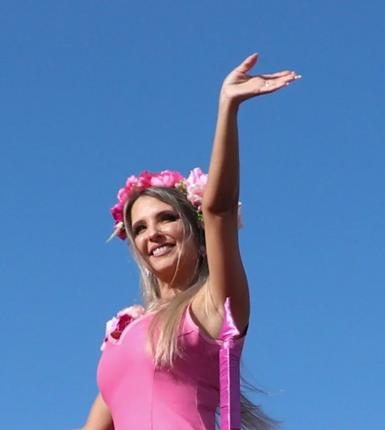 Young woman wearing a pink dress and flower crown, smiling and raising her arm against a clear blue sky.