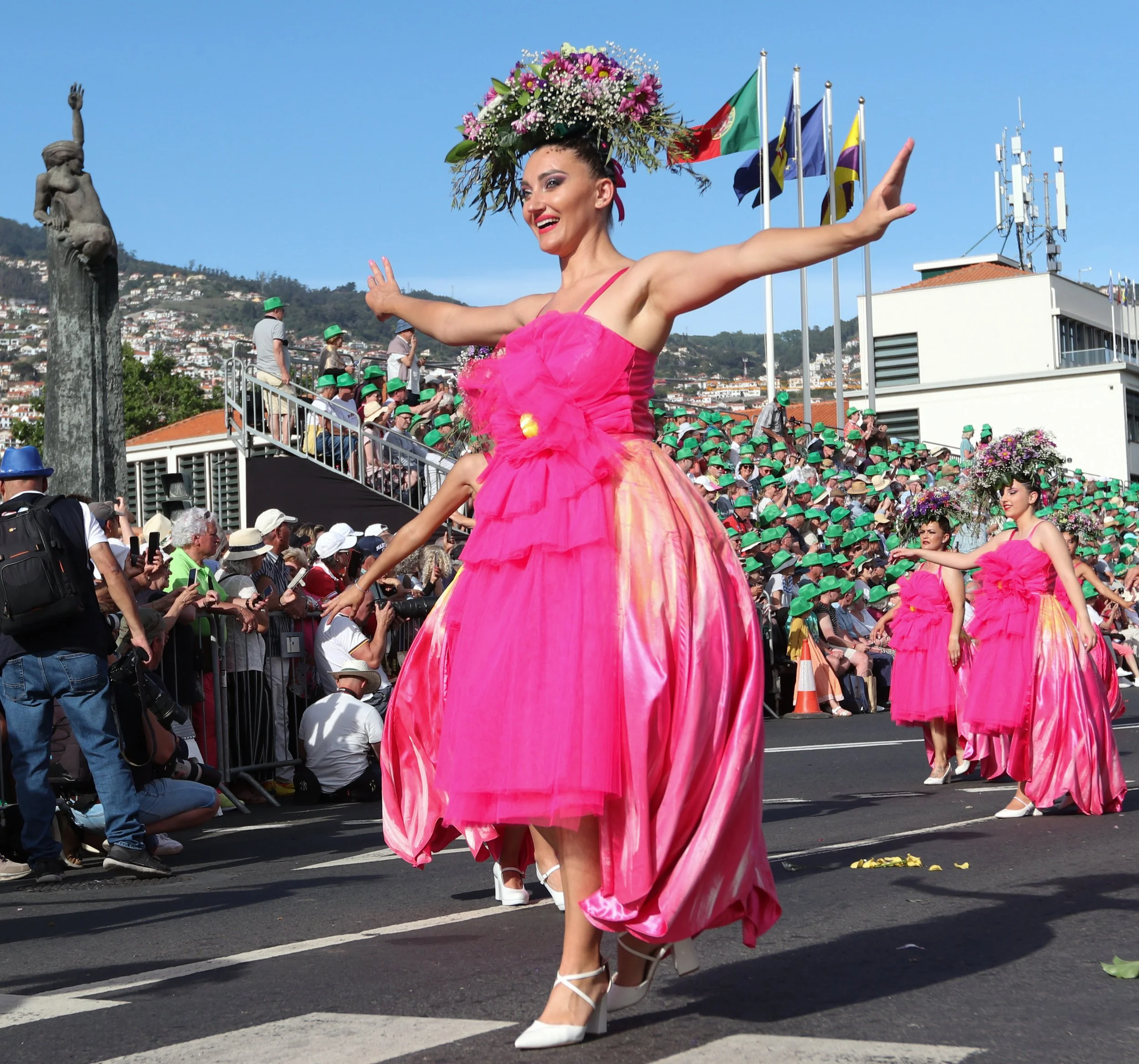 Women in pink costumes performing a parade, with a woman in the front dancing with arms outstretched, wearing a floral headpiece, and a crowd watching from the sidelines.