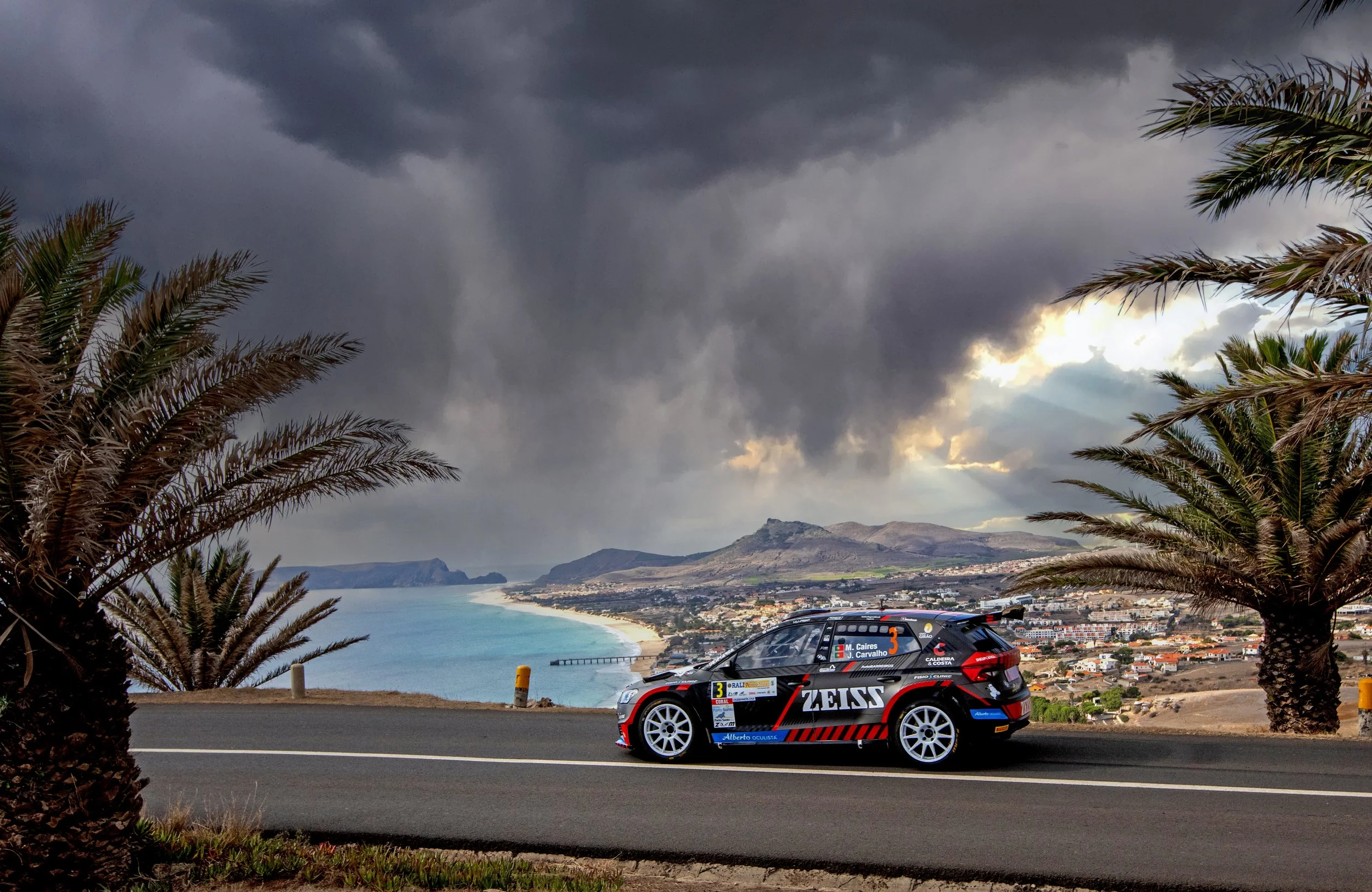 A rally car with Zeiss branding driving on a coastal road with palm trees and mountains in the background under dark, stormy clouds.