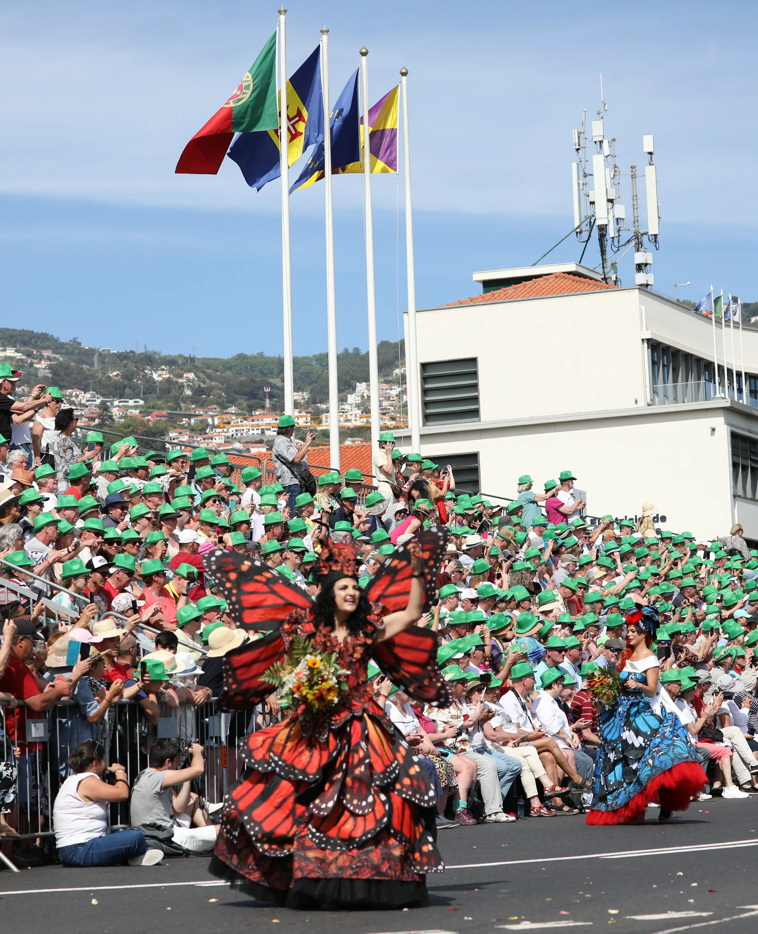 A parade with a large crowd of people wearing green hats, two women dressed in butterfly costumes with large orange and black wings, one holding flowers, are walking along the street, with flags flying overhead and a building with antennas in the bac