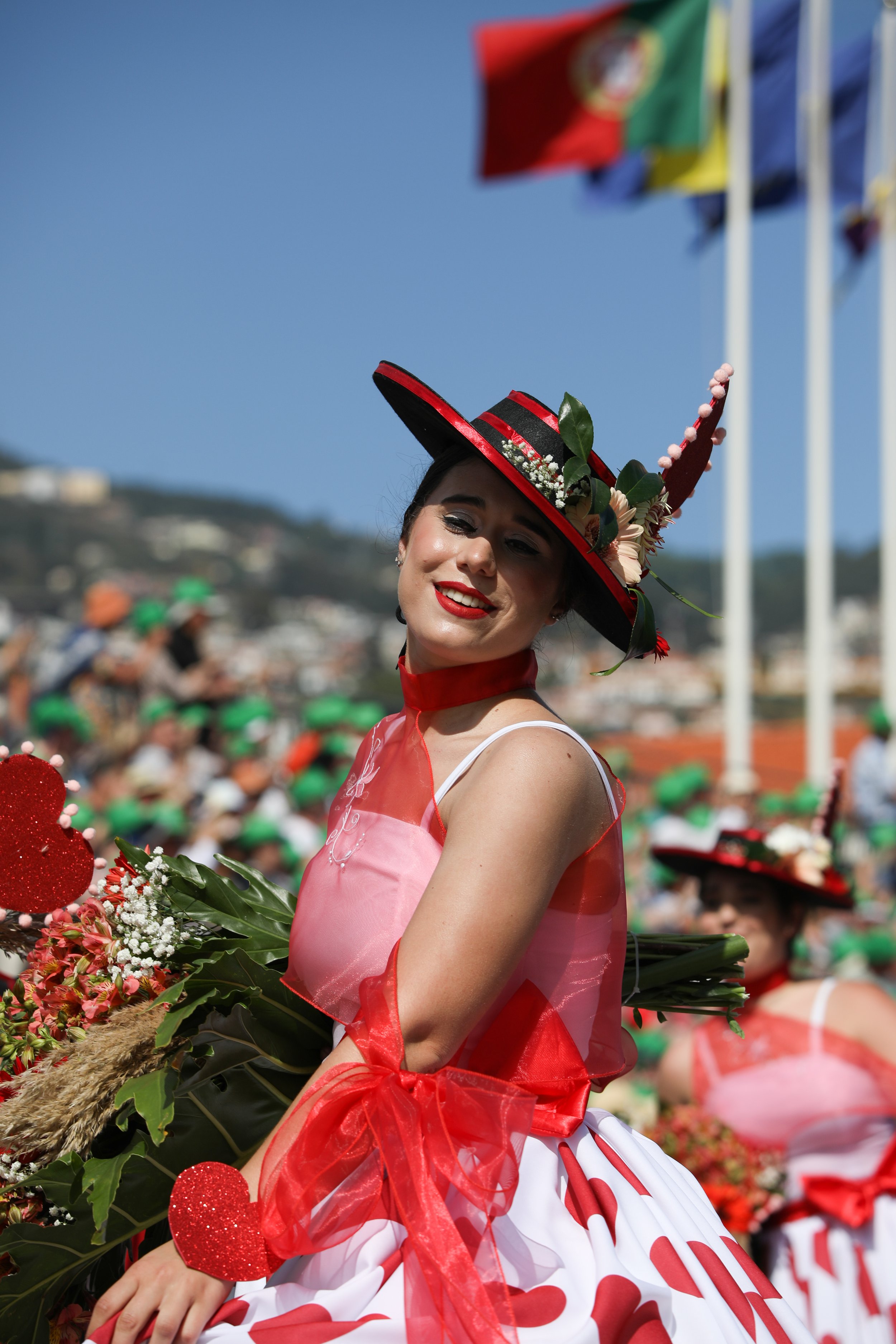 A woman dressed in traditional Mexican attire, holding flowers, and wearing a hat with decorations, participating in a cultural parade or celebration outdoors on a sunny day.