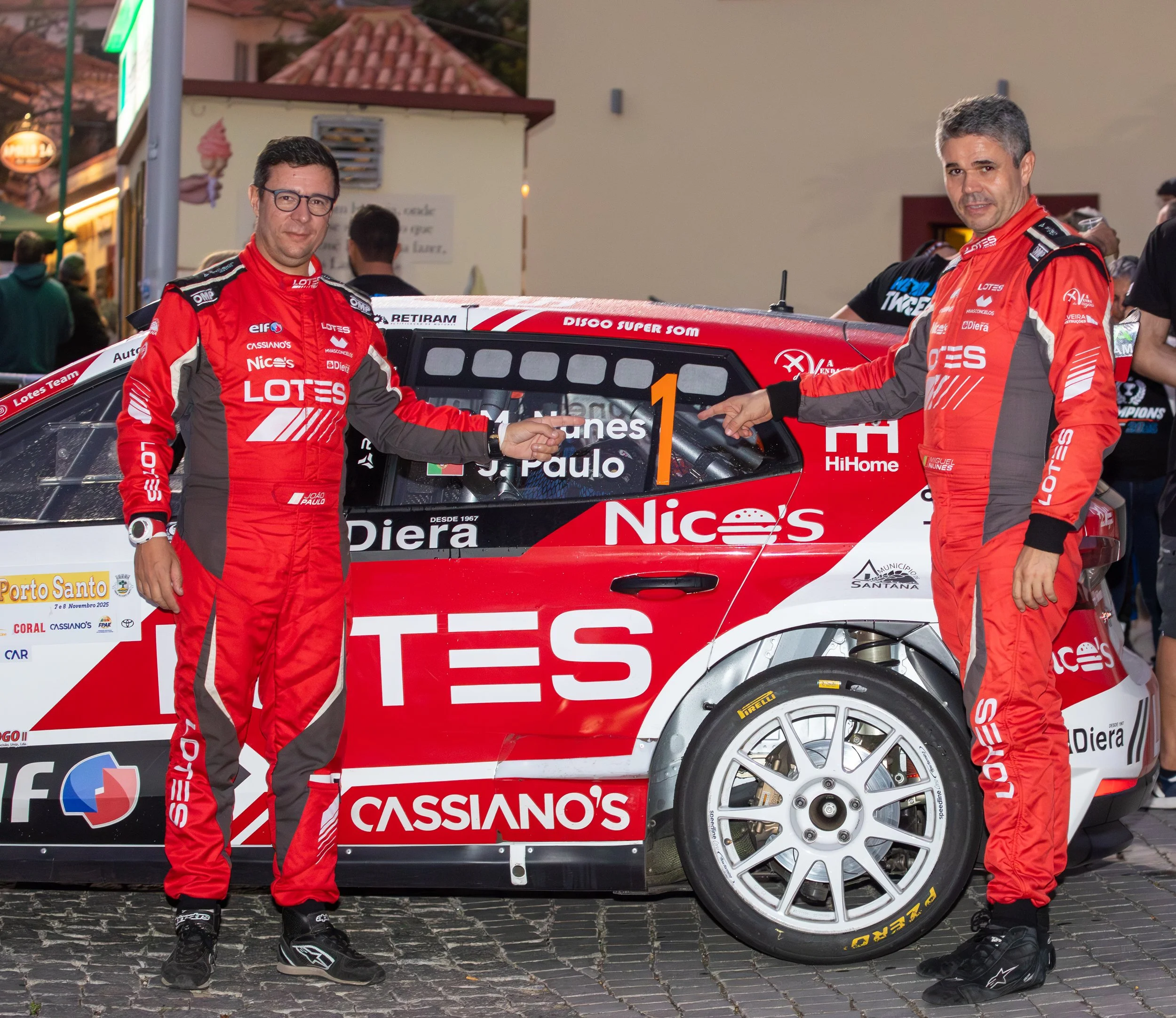 Two race car drivers in red racing suits standing next to a red and white race car, with one person pointing at the car. The car is decorated with various sponsor logos and the number 1.