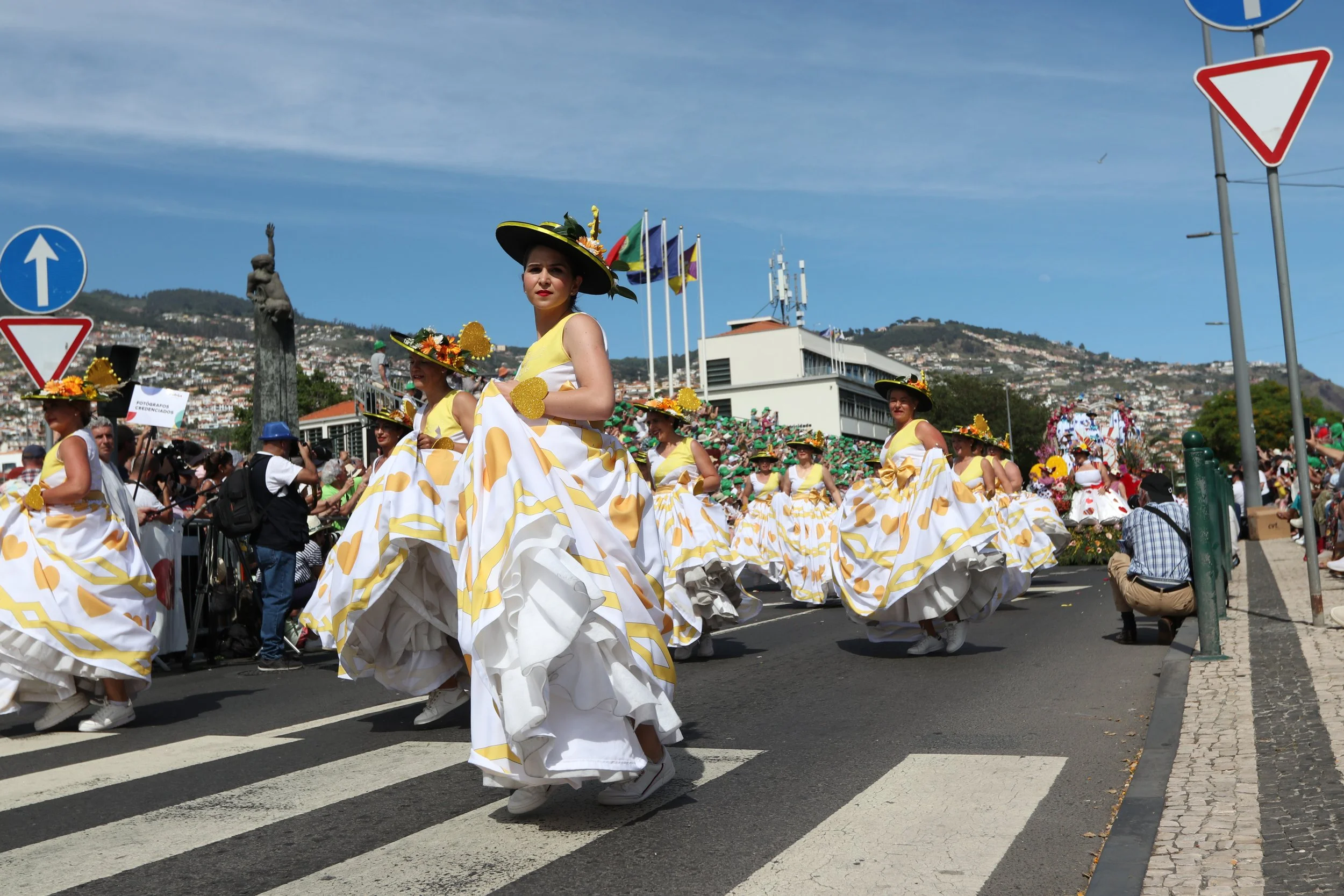 Women dressed in traditional yellow and white dresses with large skirts and hats with flowers participating in a parade on a sunny day with a crowd and flags in the background.