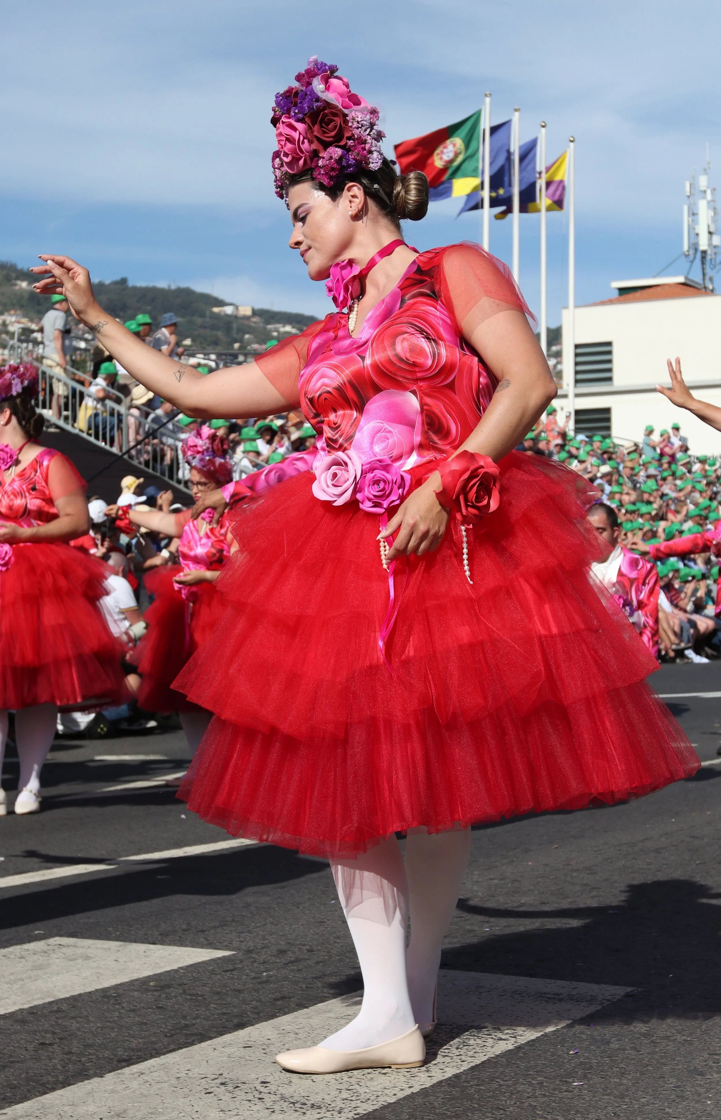 A woman dressed in a vibrant red and pink floral costume with a tulle skirt, floral headpiece, and pearl accessories, participating in a parade.