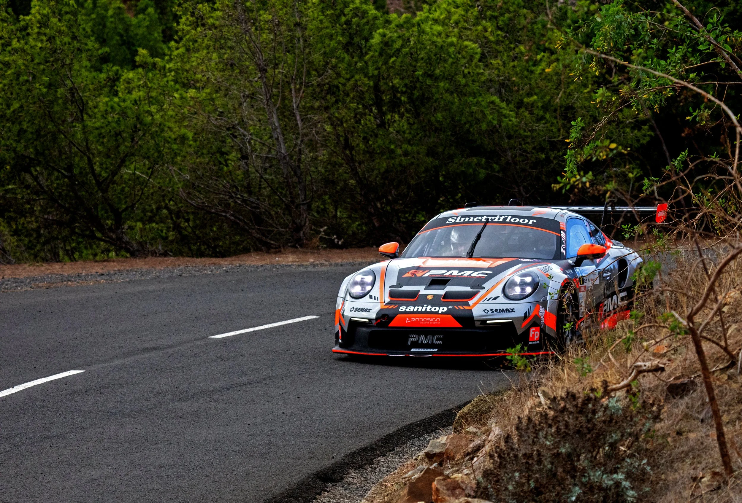 A race car on a winding road surrounded by trees and bushes, with various sponsor logos and a black and orange color scheme.