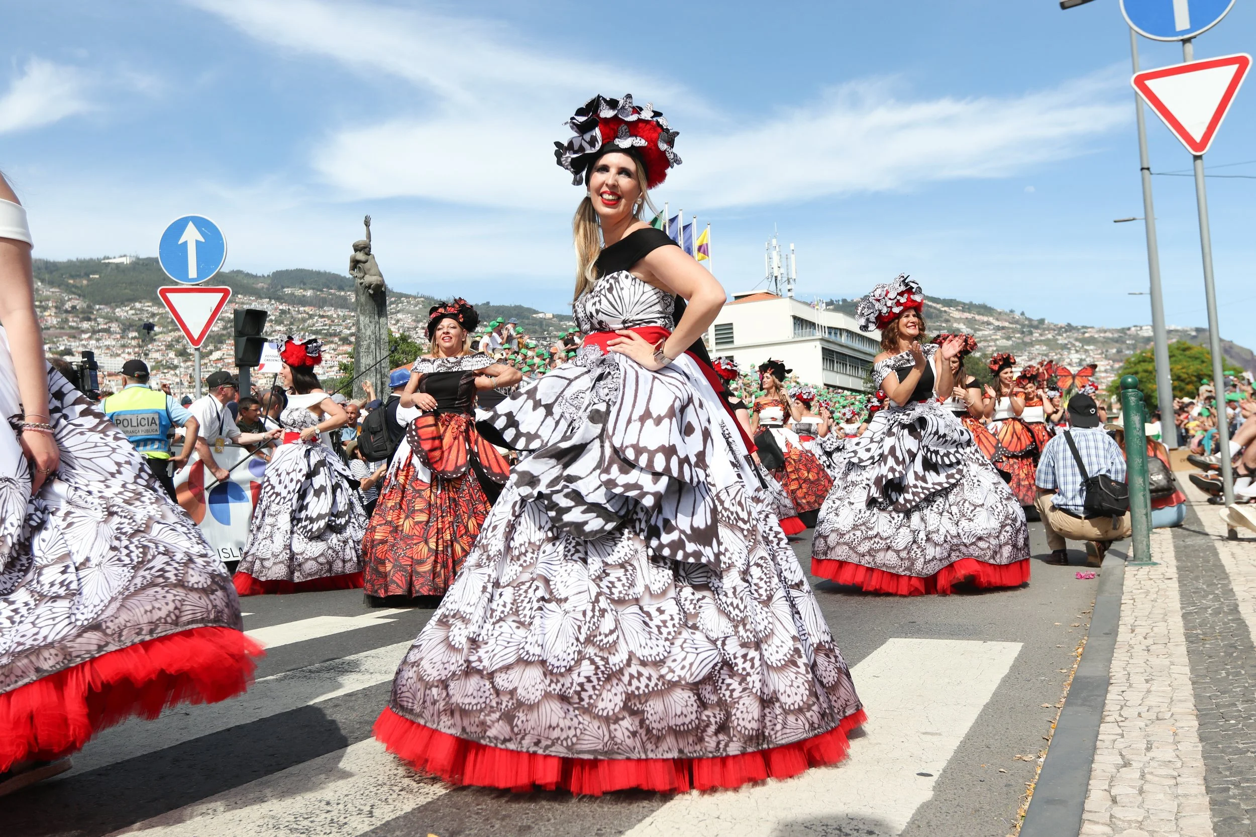 Women in colorful butterfly-themed dresses dancing in a street parade, with onlookers and police officers nearby, under a blue sky with some clouds.