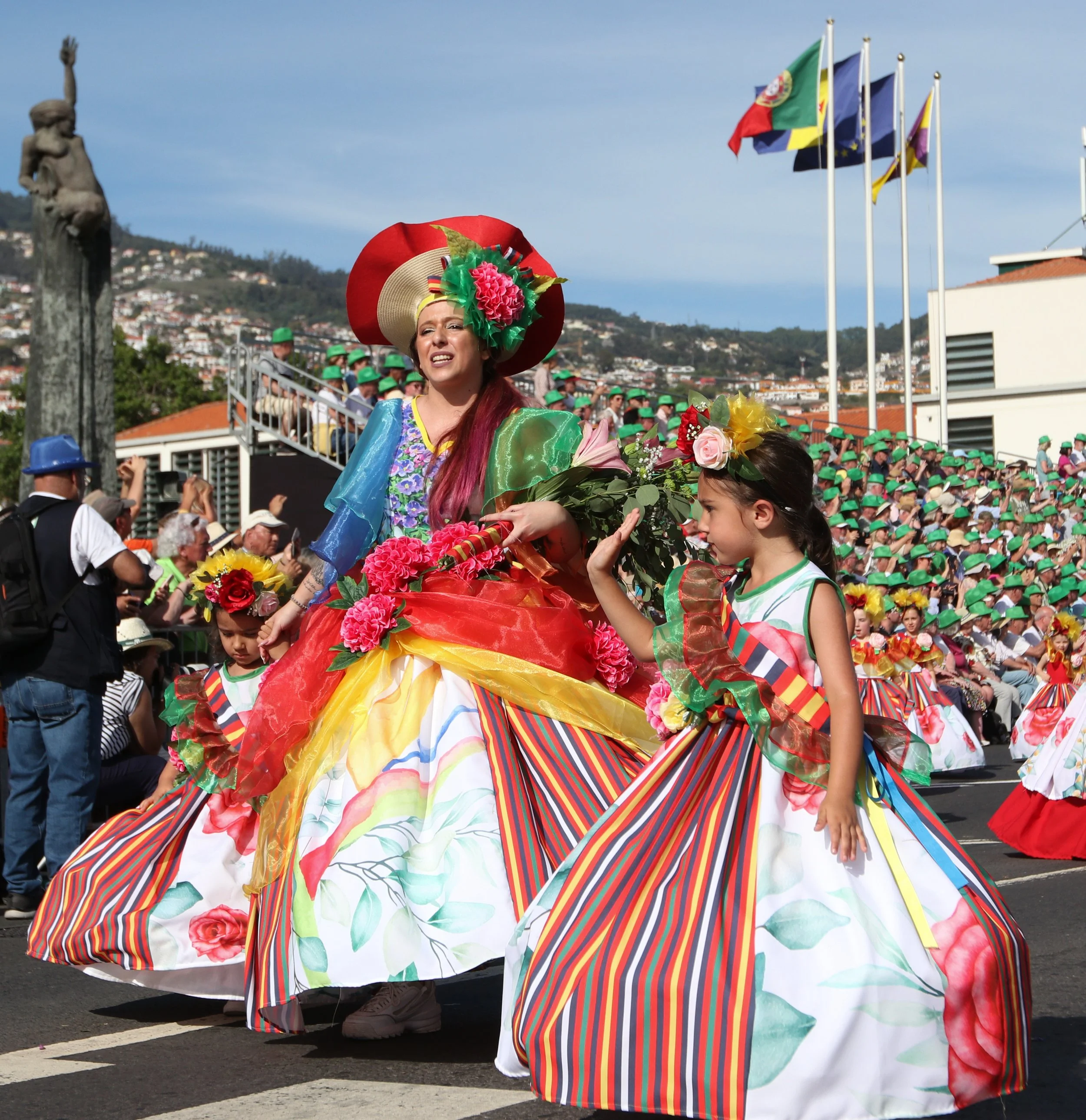 Women and children in colorful traditional costumes parade on a city street during a cultural festival, with spectators and flags in the background.