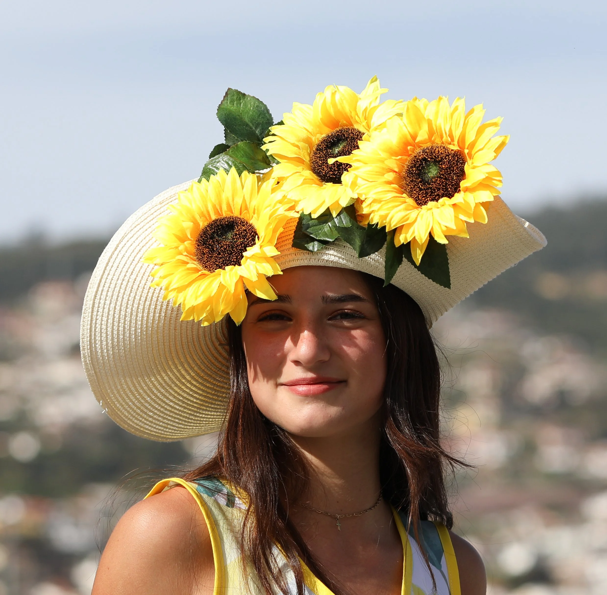 Young woman wearing a large sun hat decorated with bright yellow sunflowers, standing outdoors on a sunny day.