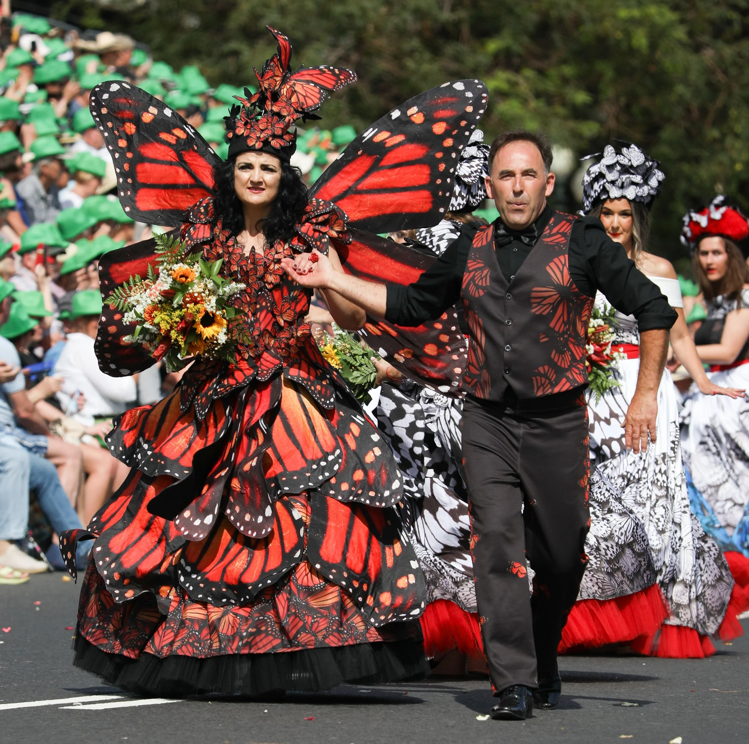 Individuals dressed in butterfly-themed costumes participating in a parade, with a woman in a large butterfly dress and matching wings holding flowers, walking alongside a man in a black shirt and vest with butterfly print, against a background of se