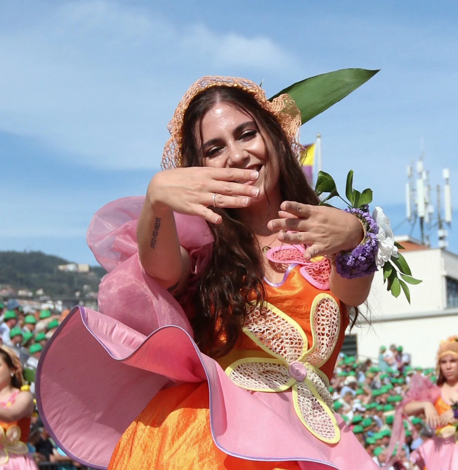 A woman dressed in a vibrant, flower-inspired costume with pink and orange hues, wearing a floral headpiece, smiling, and throwing pink flower-shaped confetti at a lively outdoor event with many people in the background.