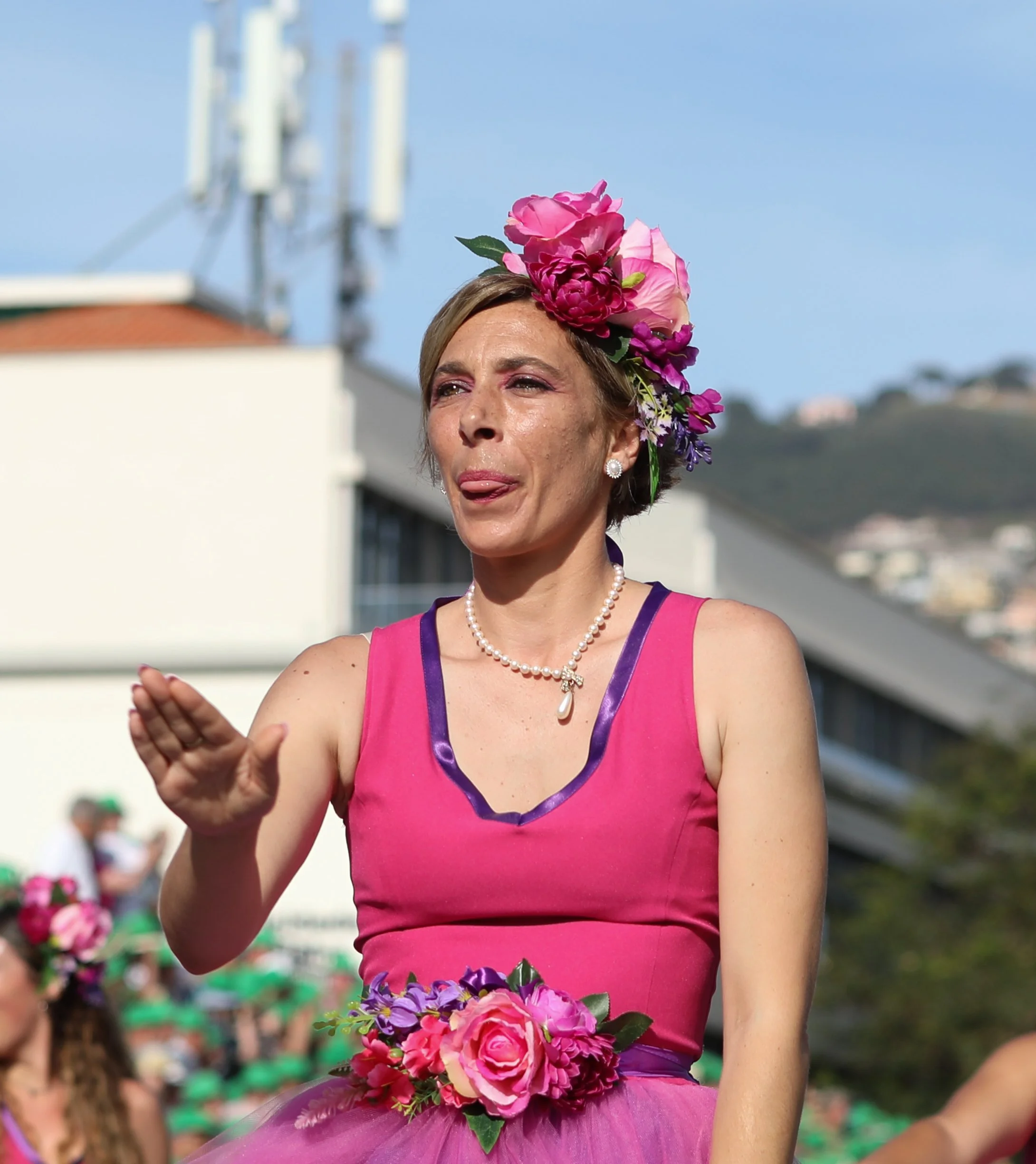 A woman wearing a pink dress with purple trim, a pearl necklace with a pendant, and a large floral headpiece, participating in a parade or festival outdoors.